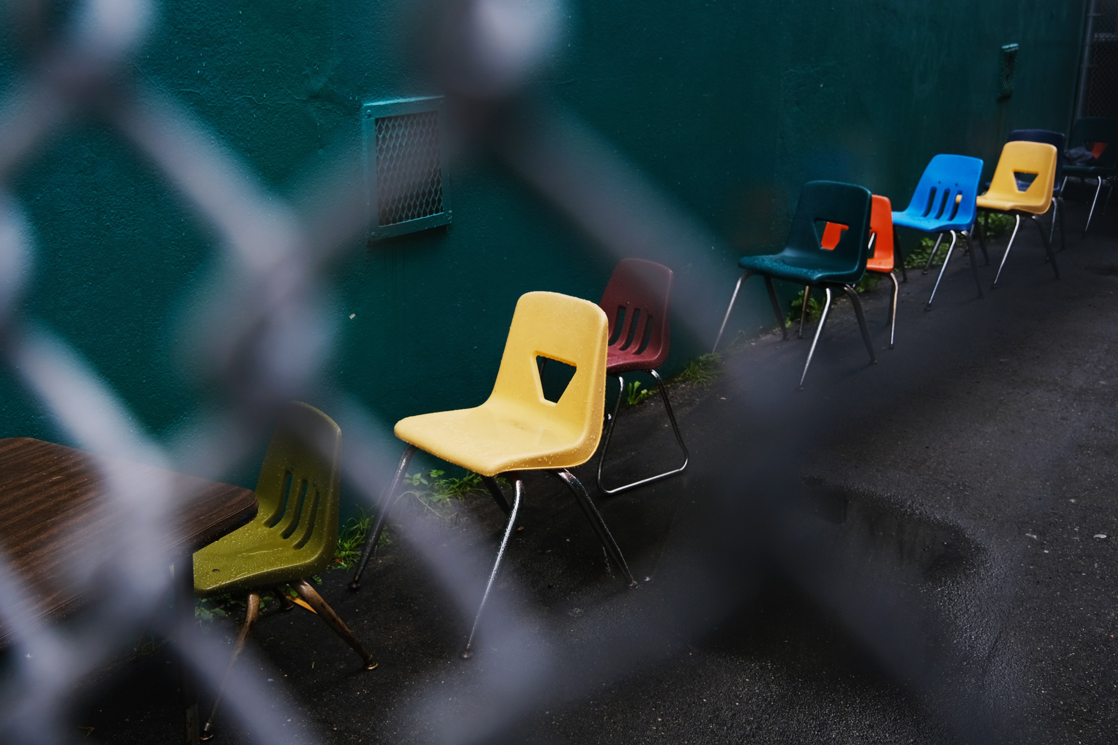 Colorful plastic chairs are lined against a turquoise wall on a wet pavement, viewed through a chain-link fence. Chairs are yellow, green, red, blue, and orange.