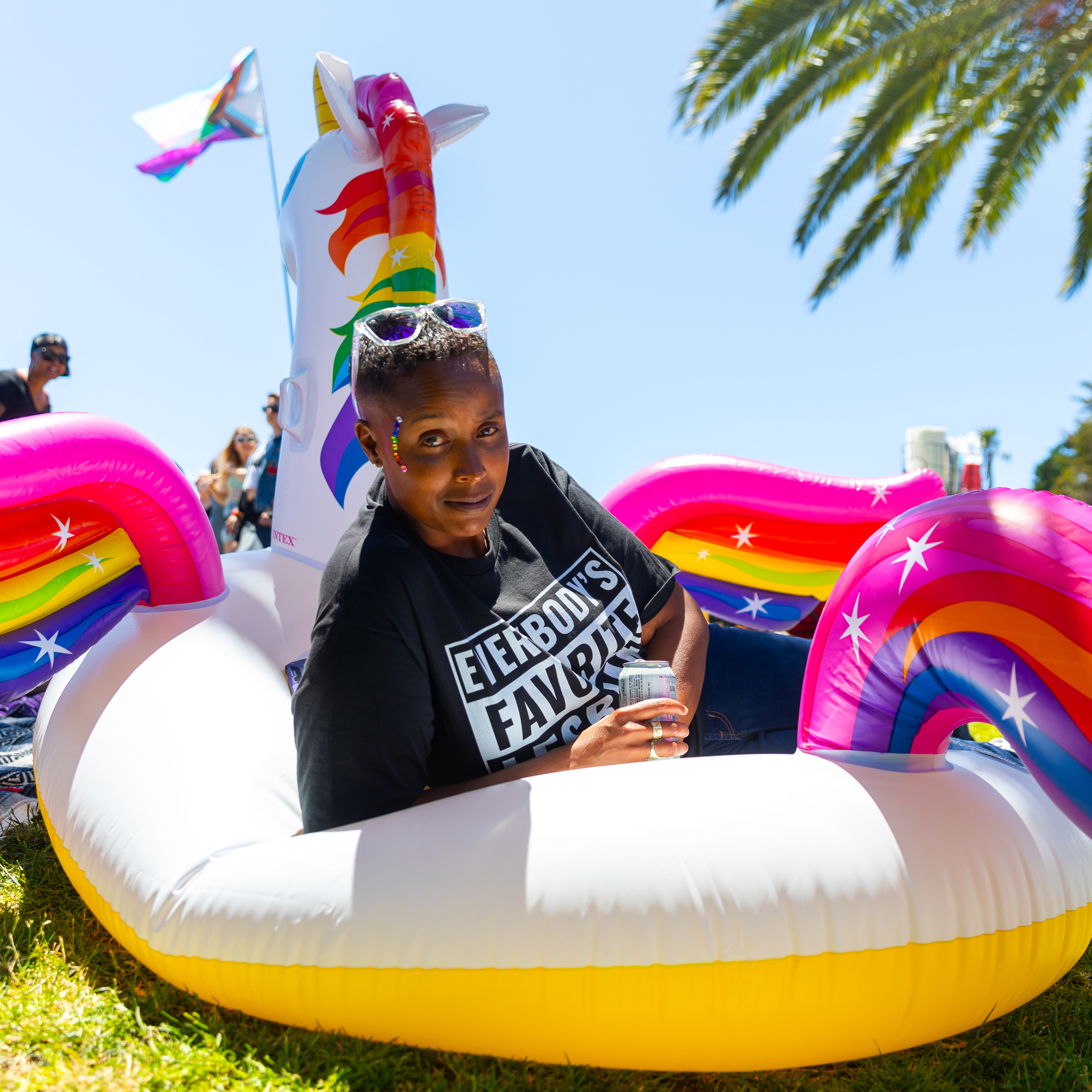 A person lounges on a colorful inflatable unicorn float, holding a drink. They wear a black shirt with white text and have purple glasses and glitter on their face.
