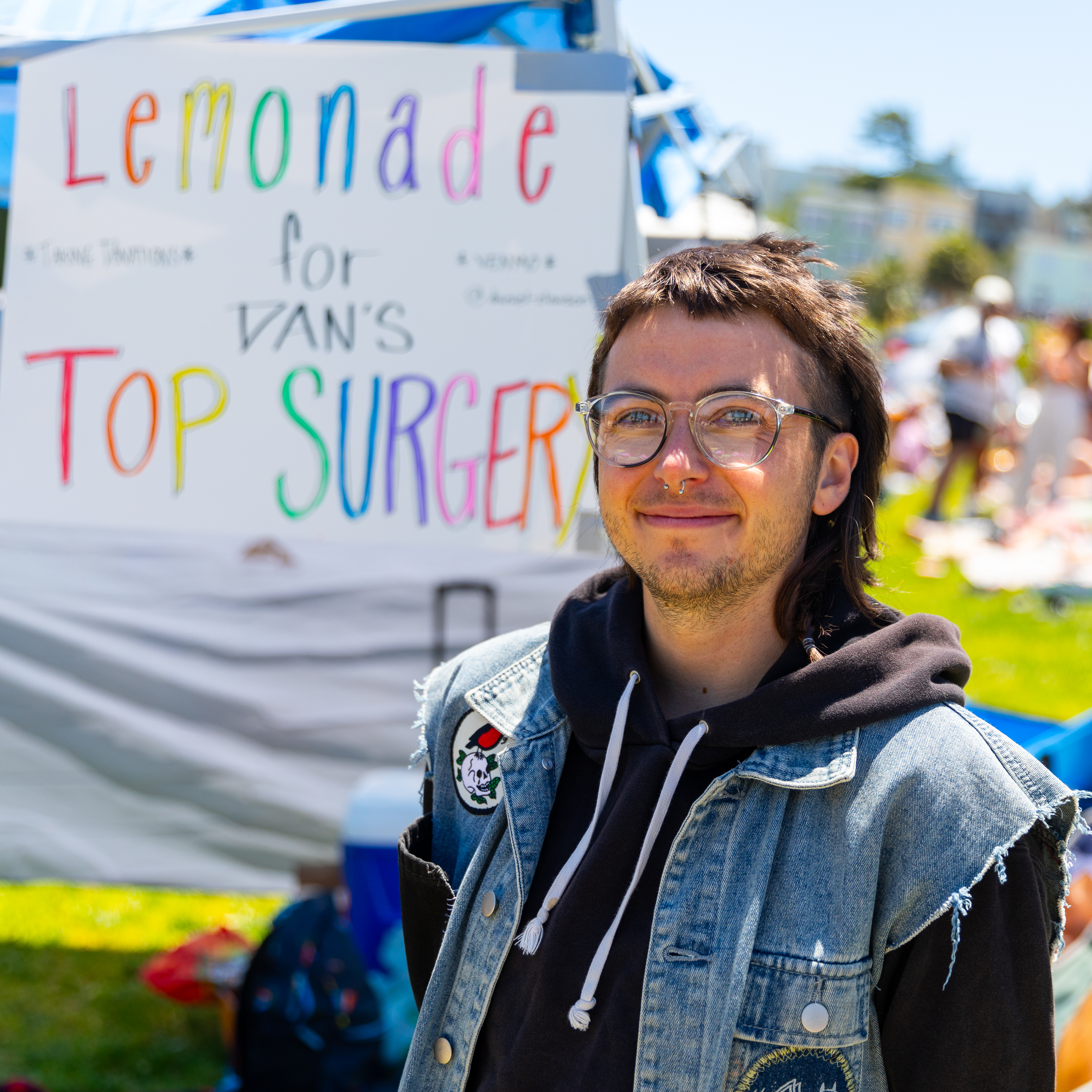 A smiling person with glasses and a denim jacket stands outdoors. Behind them, a sign reads &quot;Lemonade for Van's Top Surgery&quot; in colorful letters.