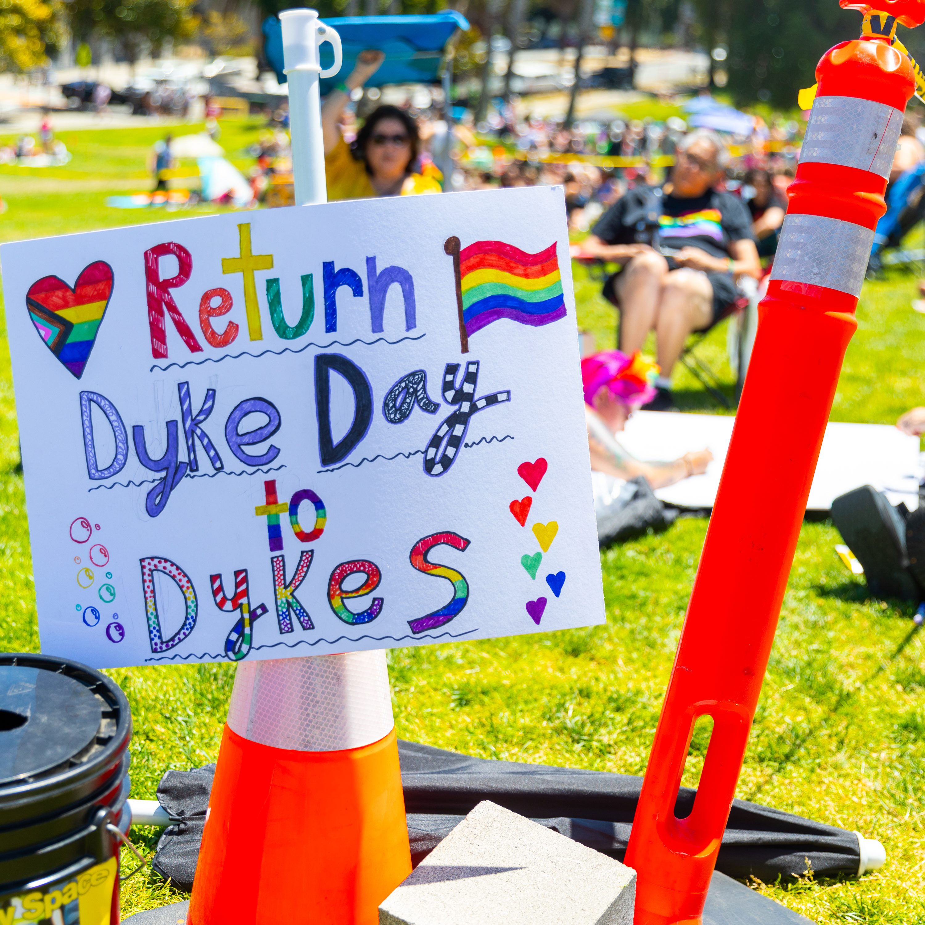 The image shows a sign with colorful text reading &quot;Return Dyke Day to Dykes&quot; with rainbow symbols, held by a cone on a grassy field with people in the background.