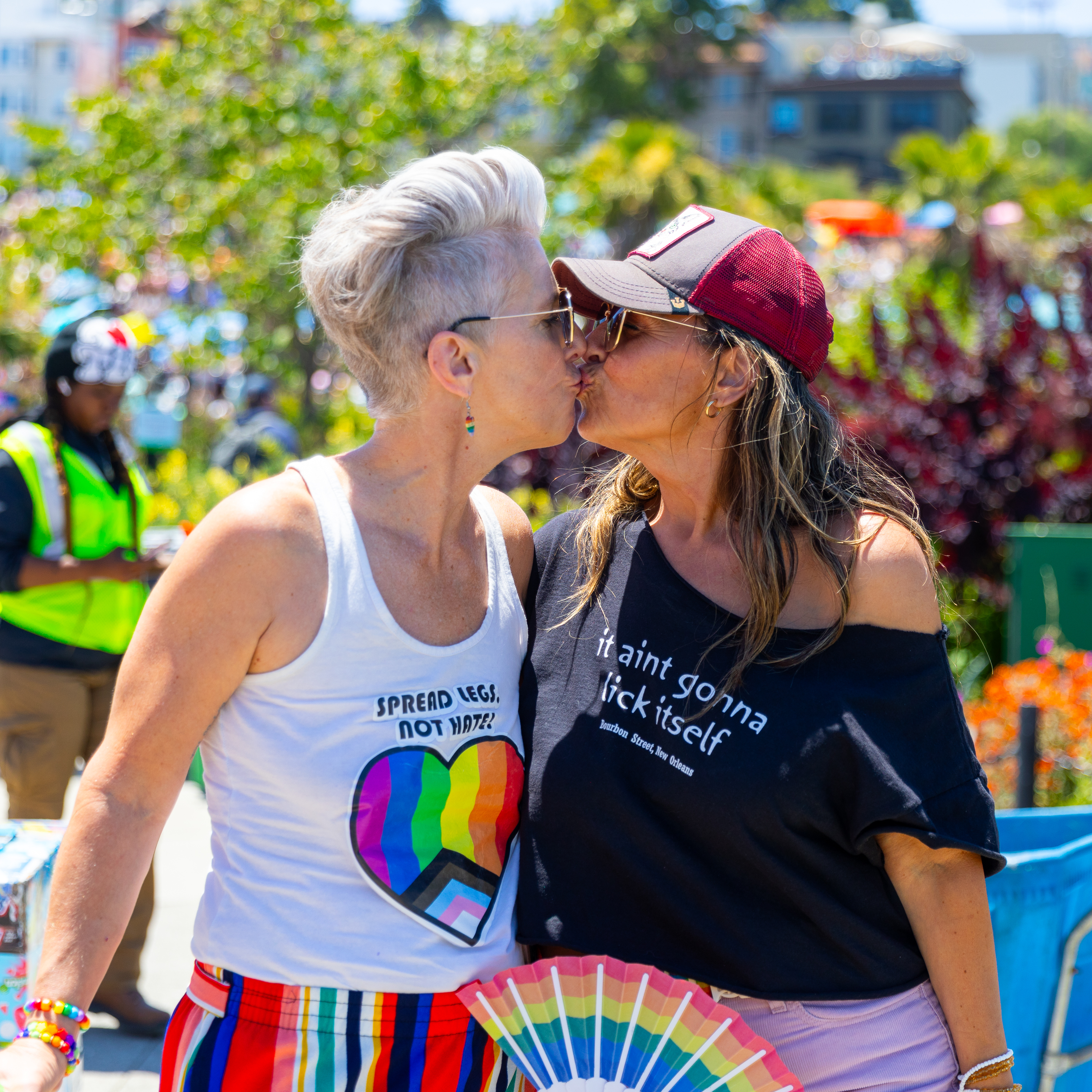 Two women wearing colorful outfits are kissing at an outdoor event. The woman on the left has short white hair, and the one on the right wears a hat and holds a rainbow fan.