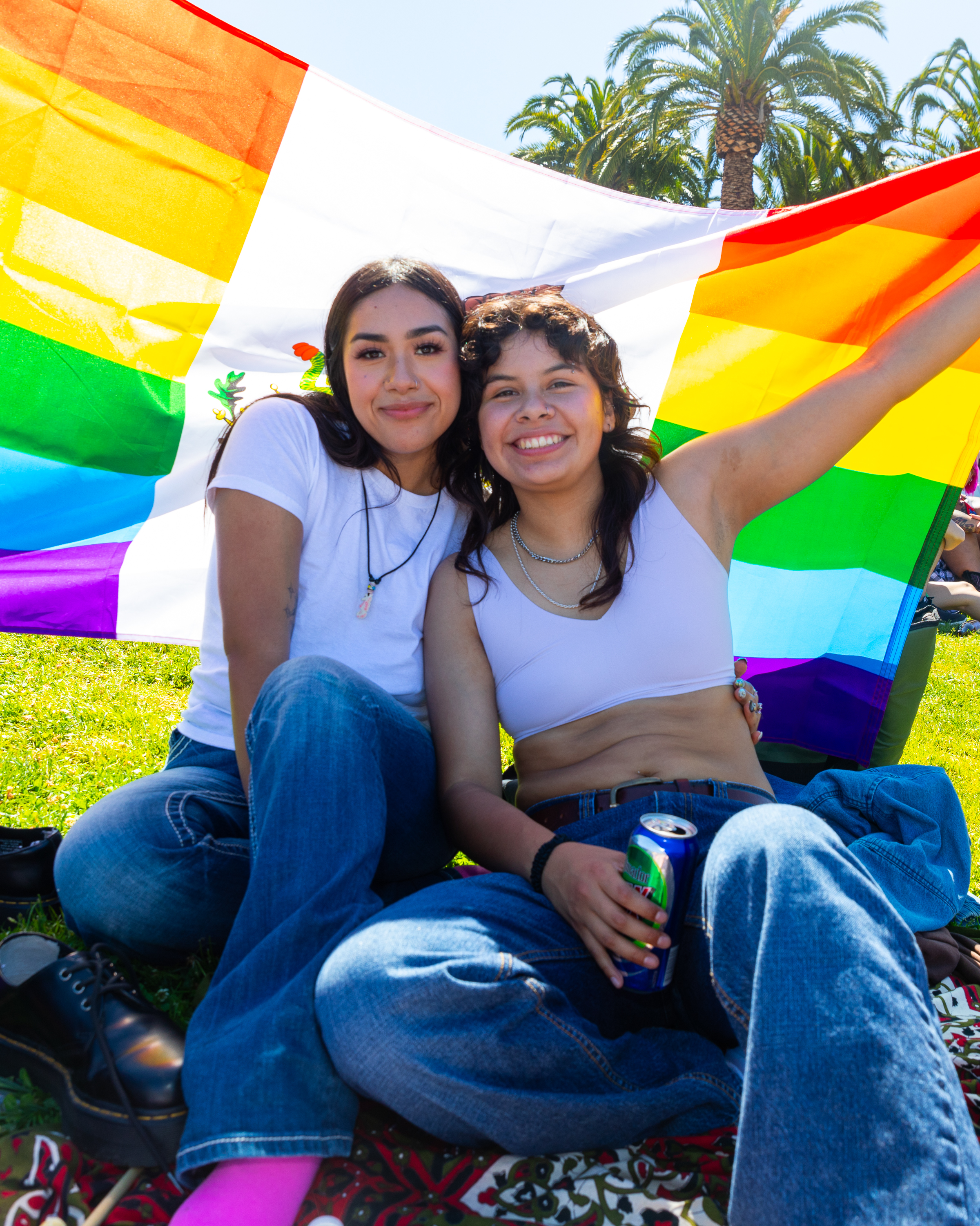 Two people sit on grass with a rainbow flag behind them, one holding a can. They are smiling, dressed casually, and palm trees are visible in the background.