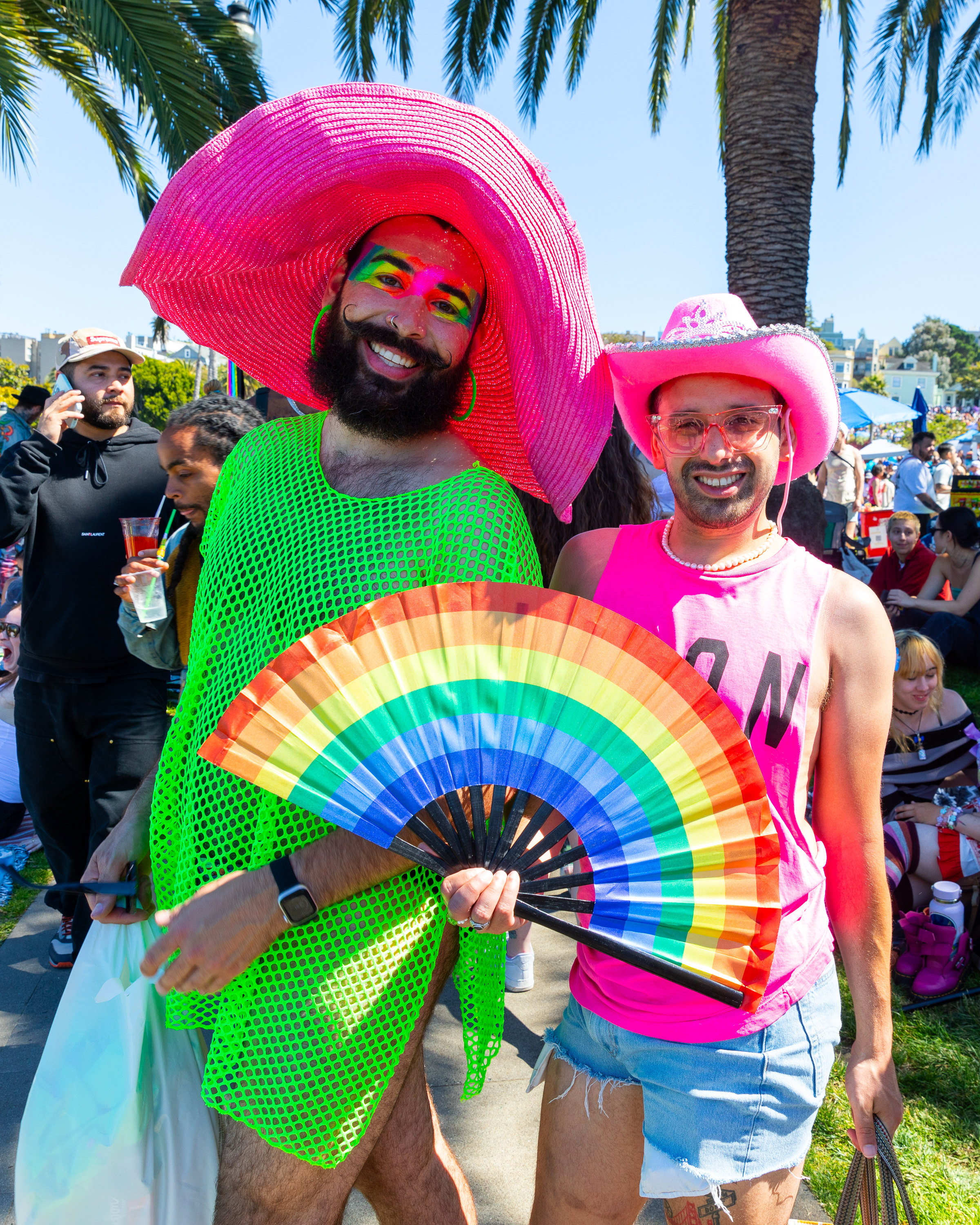 Two smiling individuals are dressed colorfully at an outdoor event beside palm trees; one in a green mesh outfit with a pink hat, and the other in pink attire and a cowboy hat.