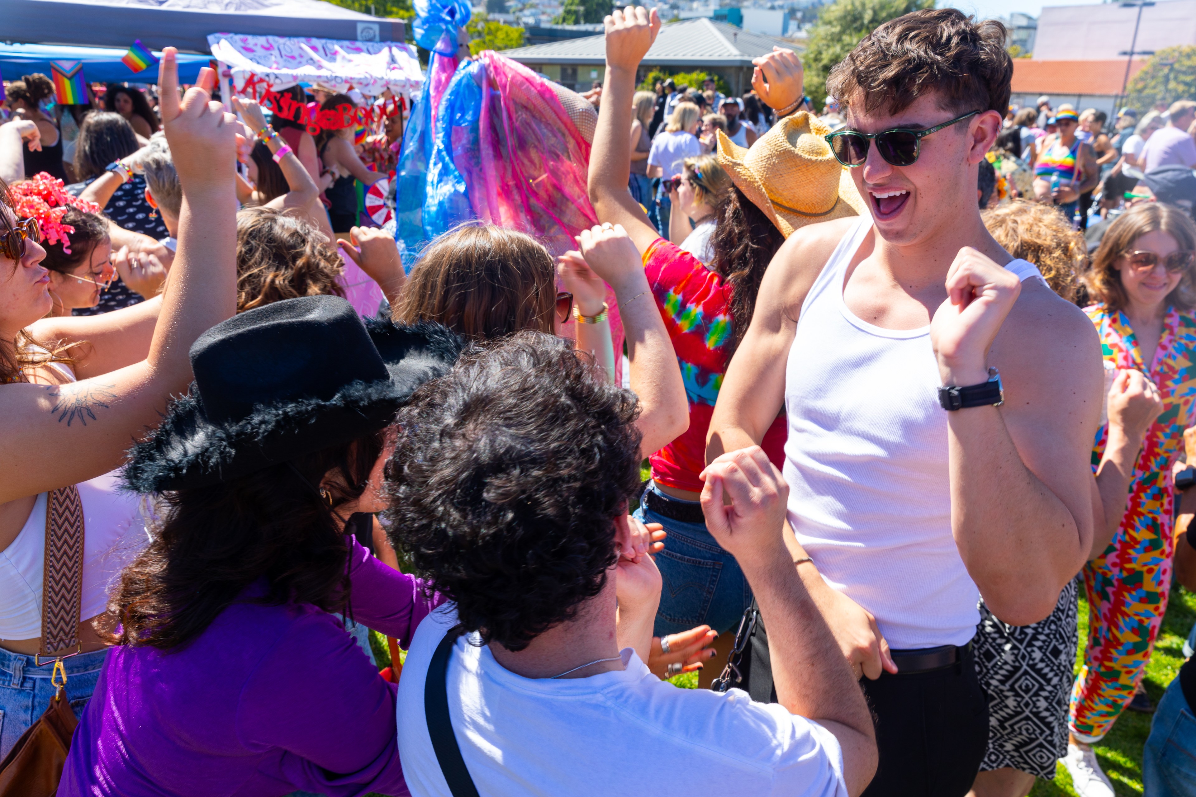 A group of people are joyfully dancing outdoors, wearing colorful outfits and sunglasses, amidst a crowd on a sunny day, creating a lively and festive atmosphere.