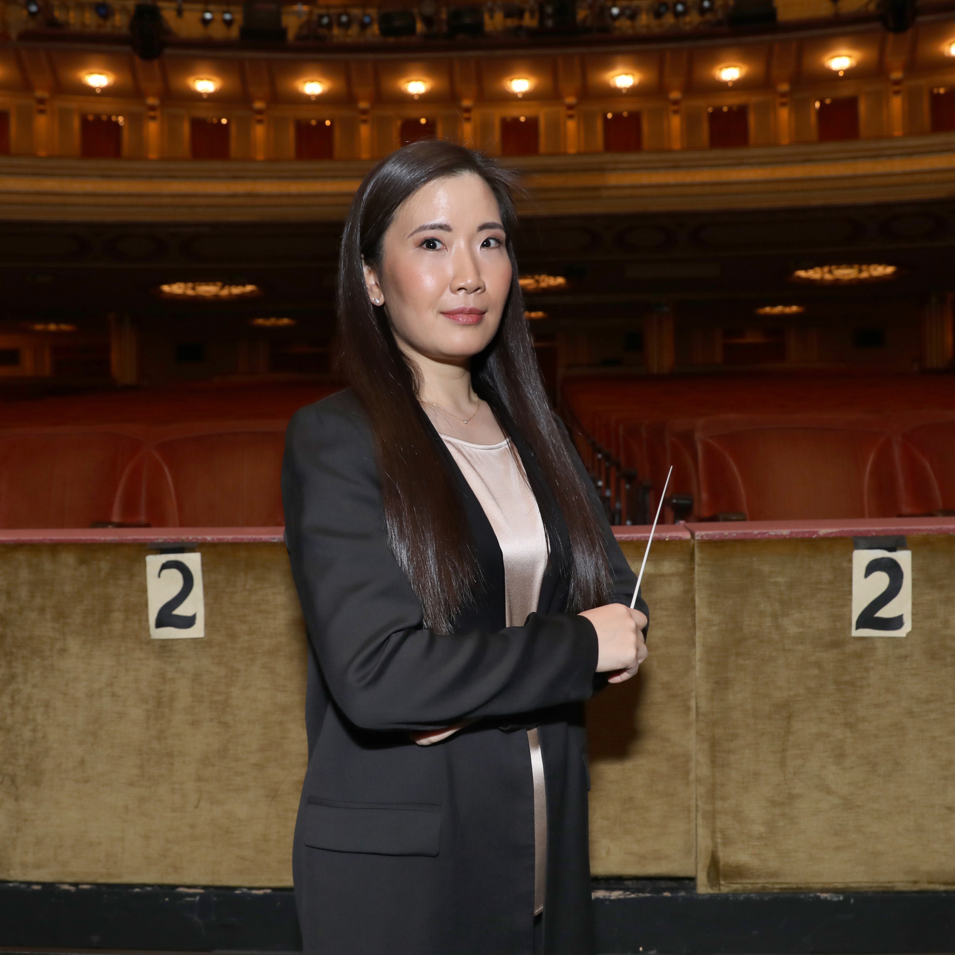 A woman with long, dark hair is standing in an empty theater, holding a conductor's baton, wearing a black blazer. The backdrop features red seats and warm lighting.
