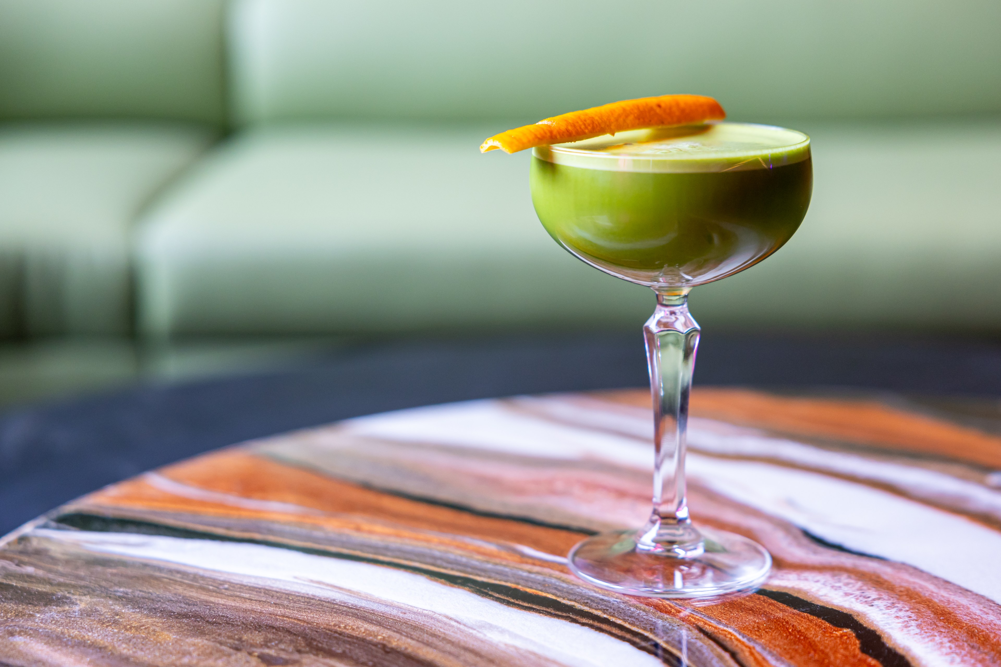 A green cocktail garnished with an orange peel in a coupe sits on a multicolored table.