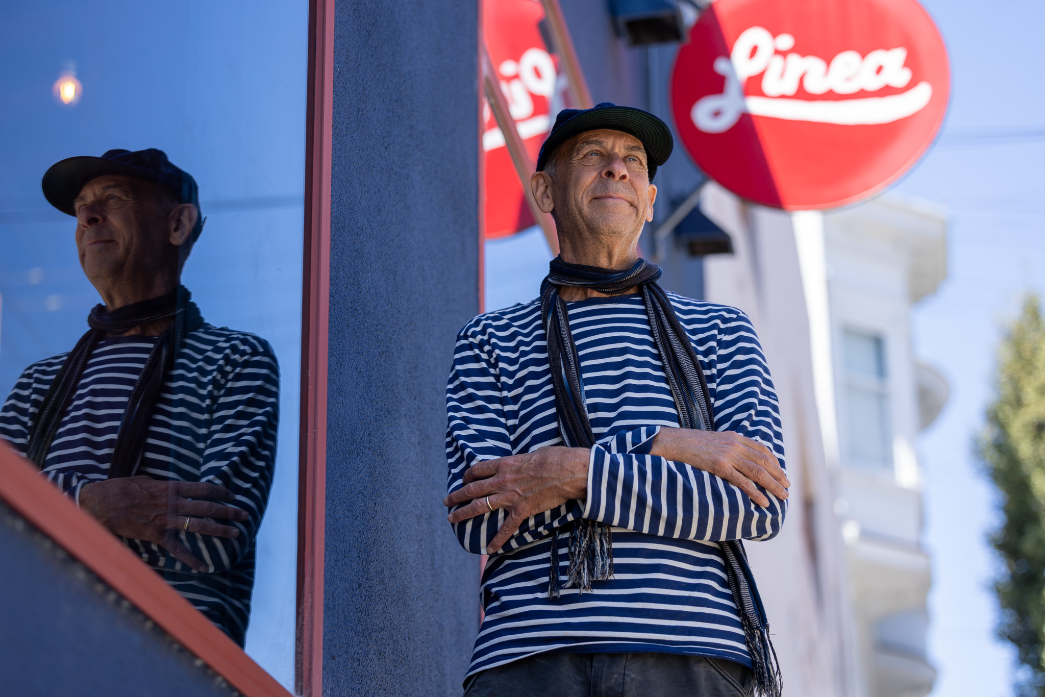 A man in a hat stands near a sign that says Linea, with his reflection in a window next to him.