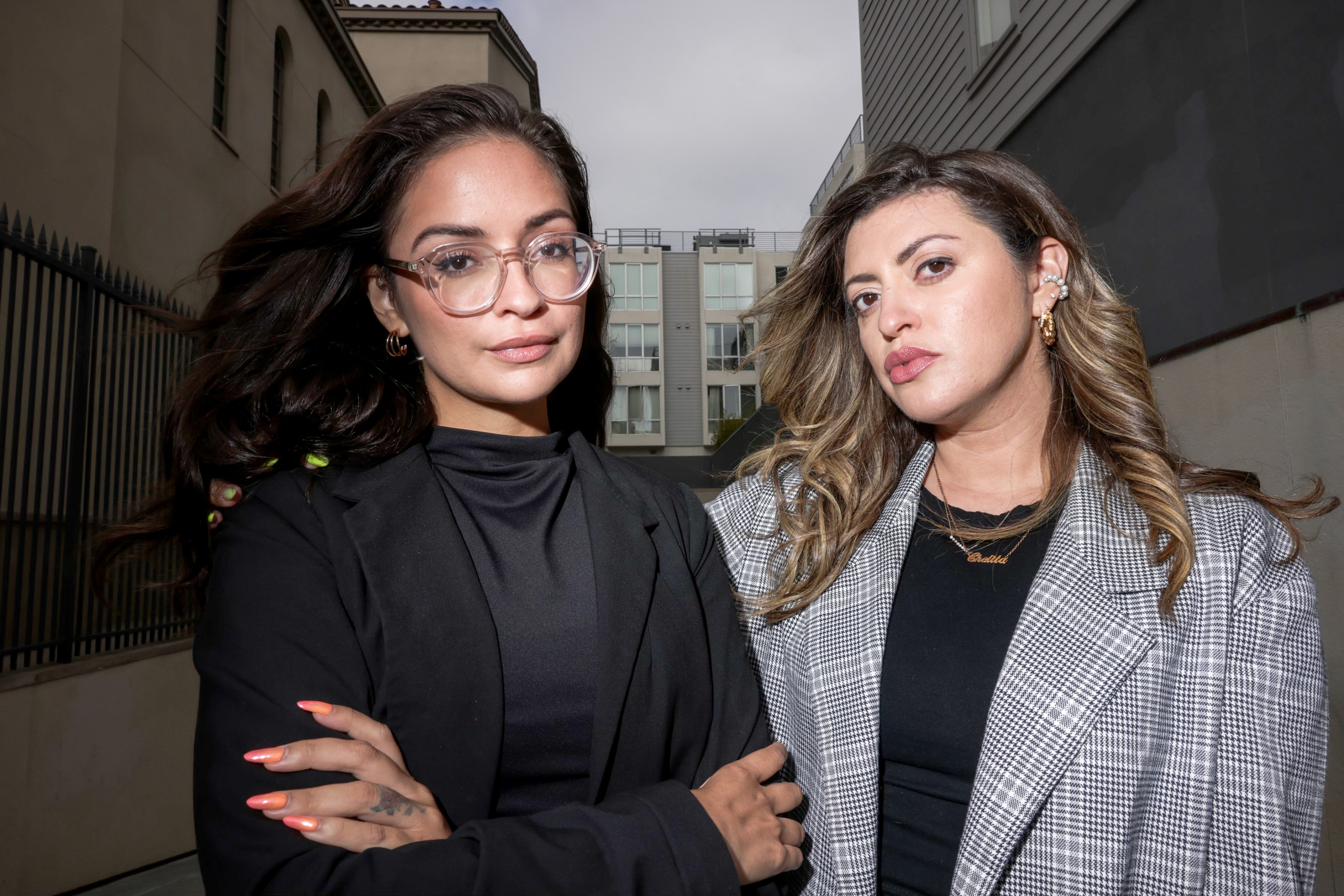 Two women stand side by side in an urban alleyway. One wears glasses and a black outfit; the other has long hair and wears a plaid blazer. Both look confidently at the camera.