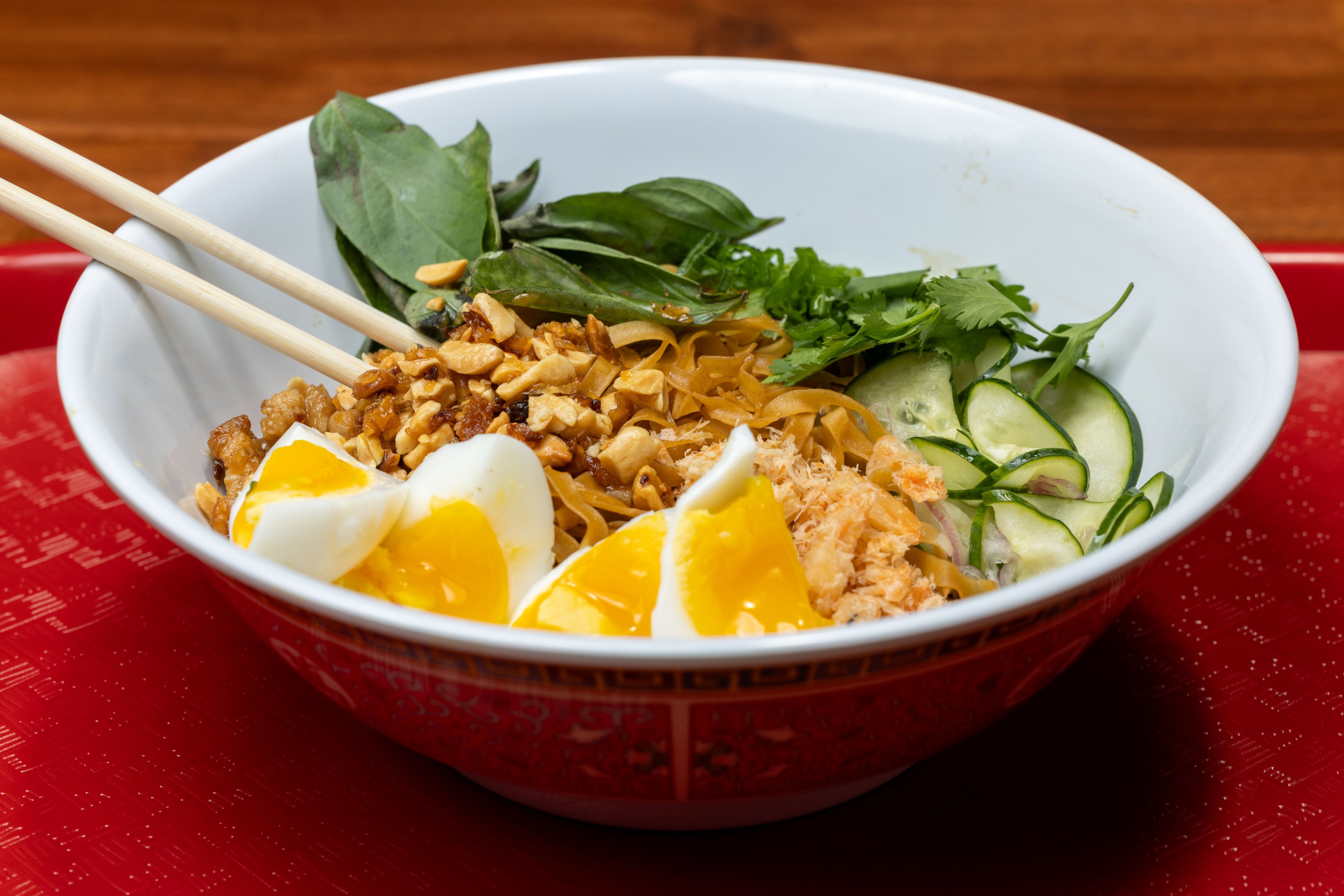 A white bowl filled with hard boiled eggs, noodles, peanuts, and fresh herbs on a red tray.