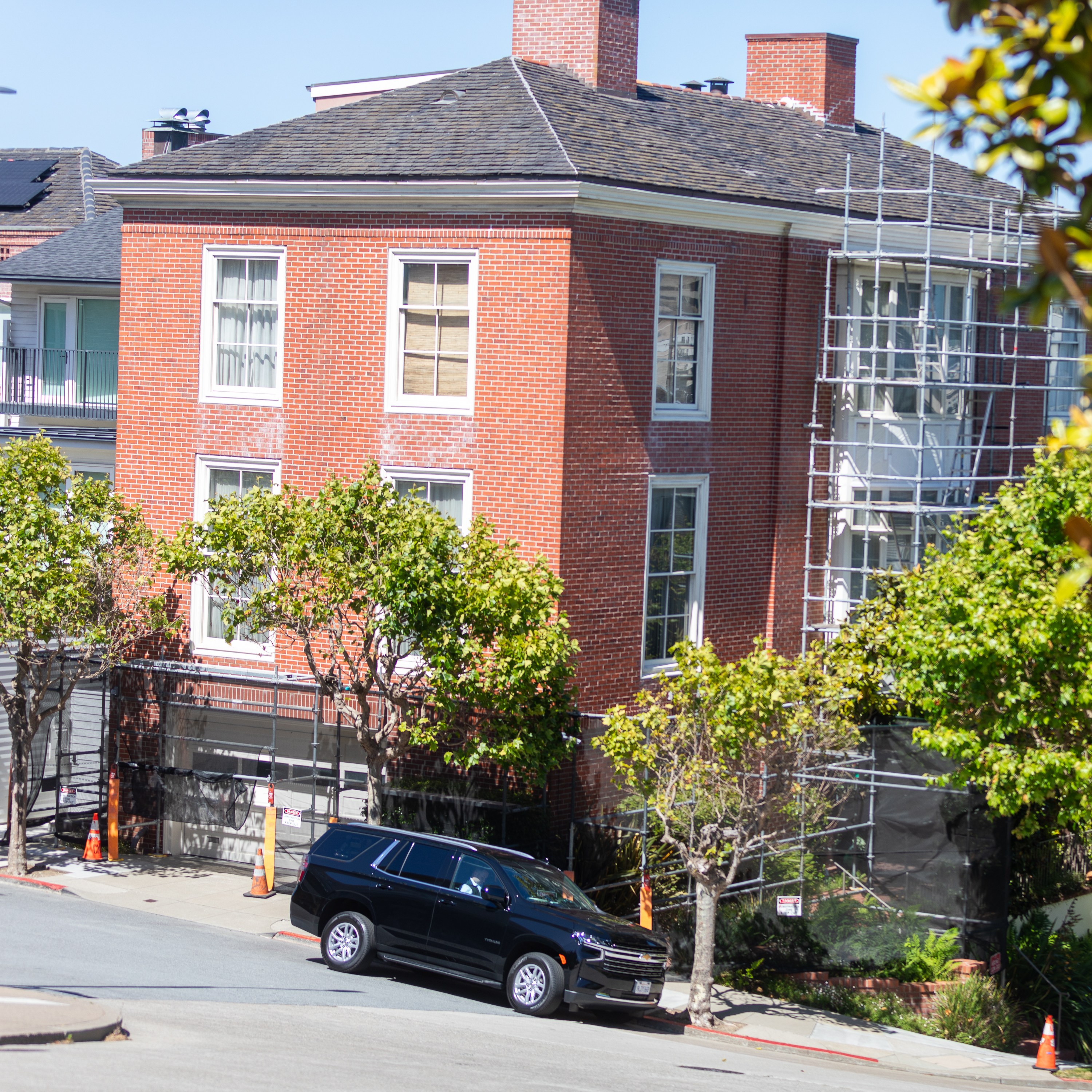 A red-brick building is undergoing maintenance with scaffolding on its side, surrounded by leafy trees and a black SUV parked in front.