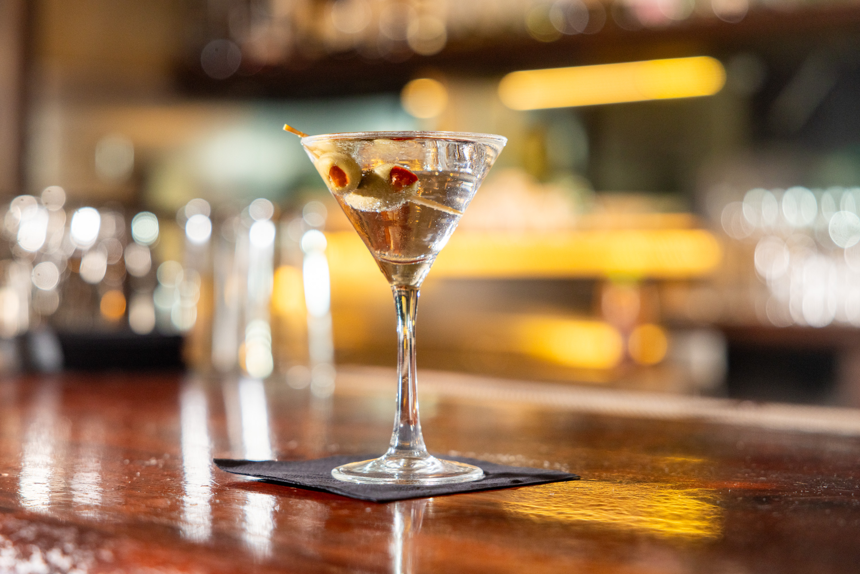 A martini glass with two olives skewered on a stick is placed on a dark square napkin on a polished wooden bar top, with a blurry background of bar lights.