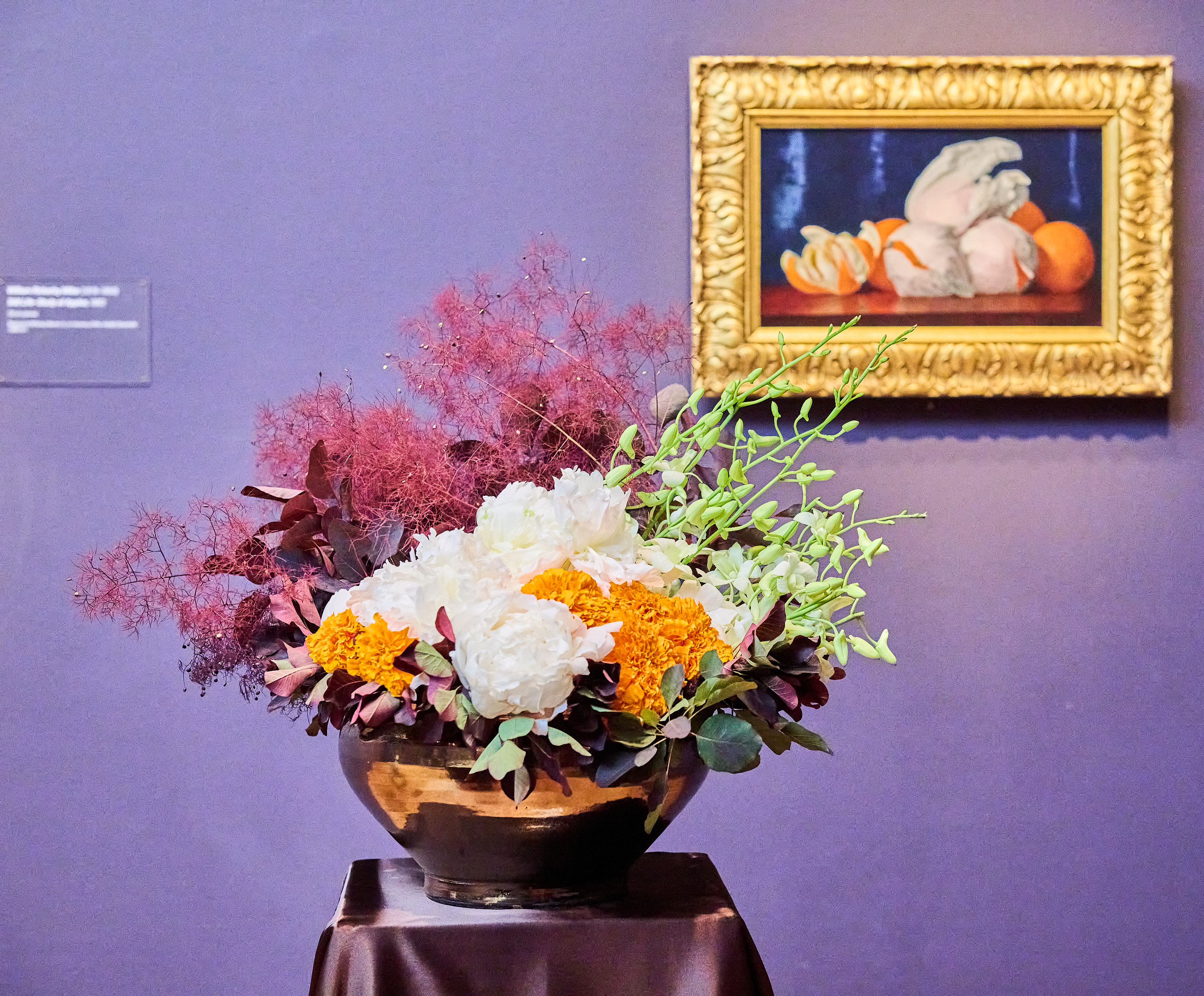 A vase with white, orange, and red flowers sits on a table with a dark cloth. A framed still-life painting of fruits hangs on the purple wall behind it.