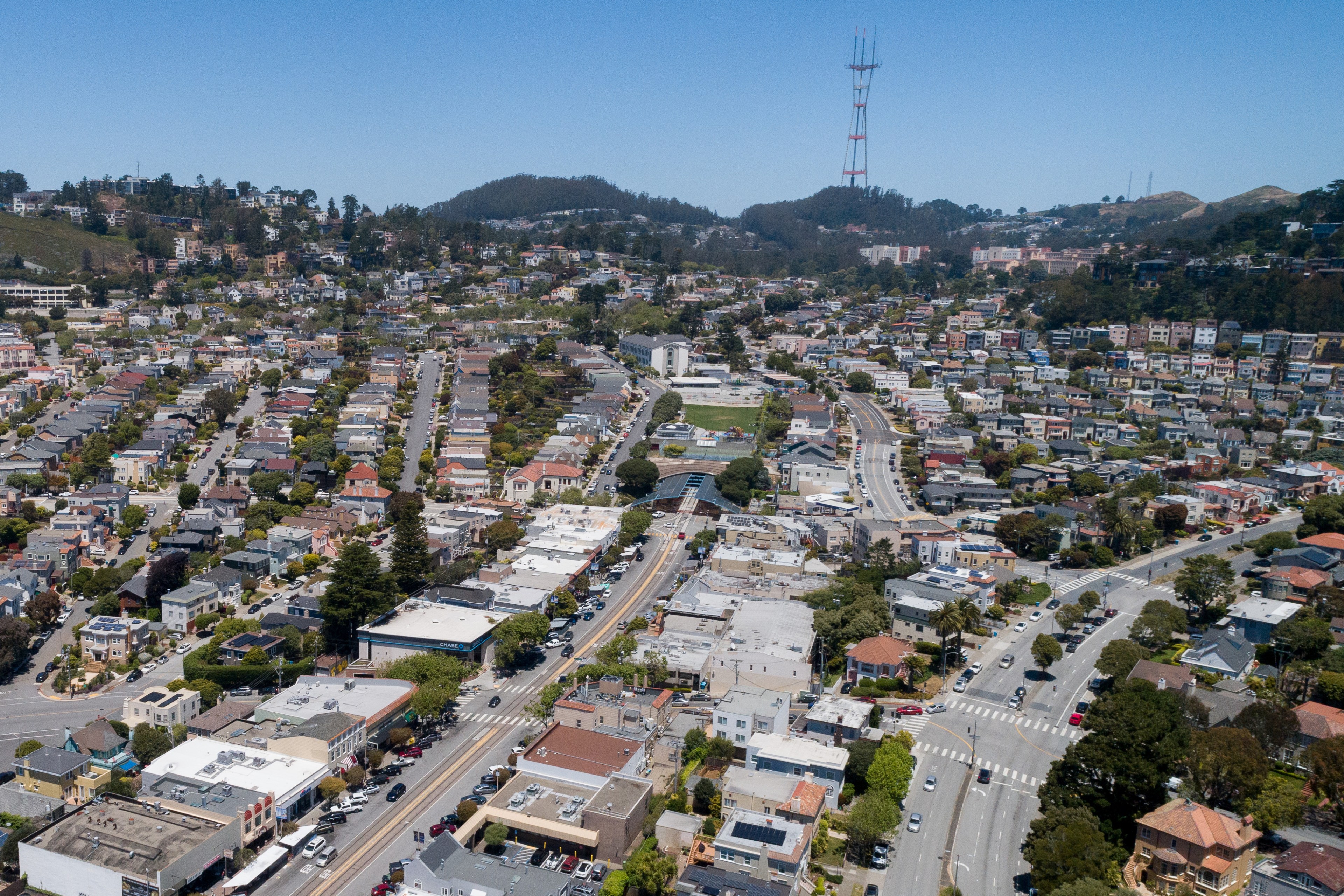 The image shows a dense urban area with rows of houses, roads, and green spaces, all set against a backdrop of hills with a tall communication tower.