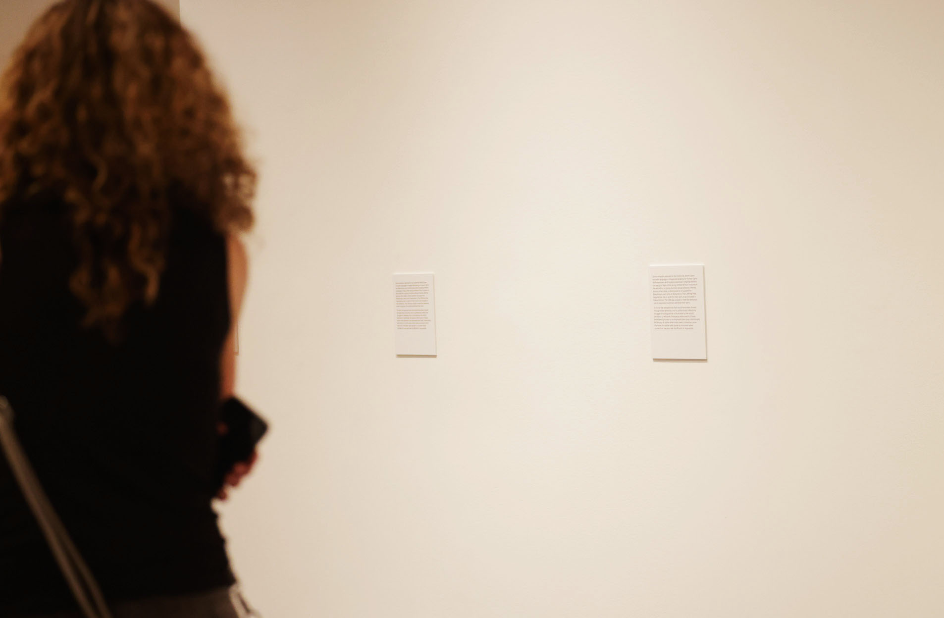 A person with curly hair, dressed in a sleeveless top, stands in front of a beige wall reading two small text panels.