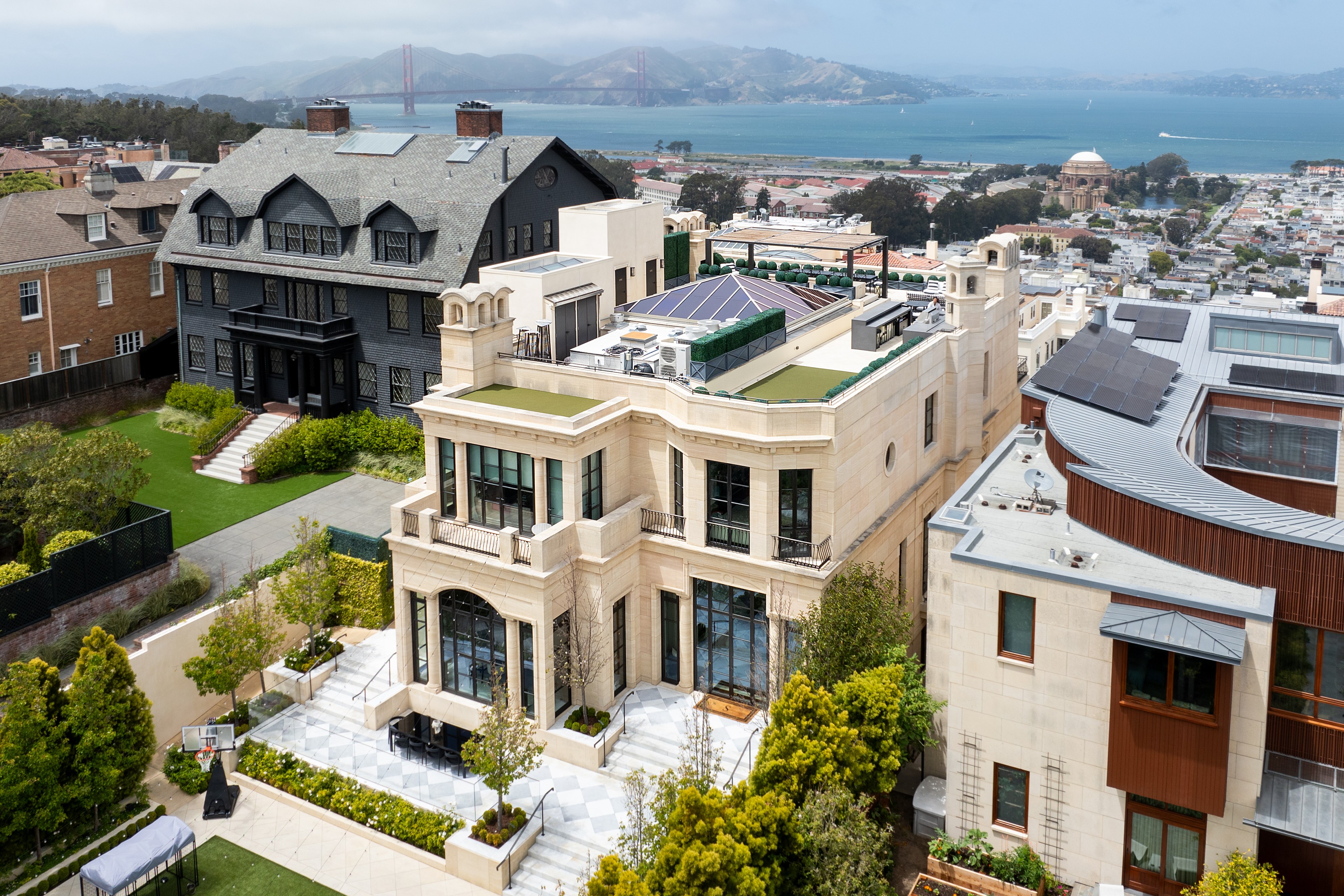 A luxurious neighborhood with elegant houses and manicured gardens. In the background, there's a scenic view of a bay with hills and a red bridge.