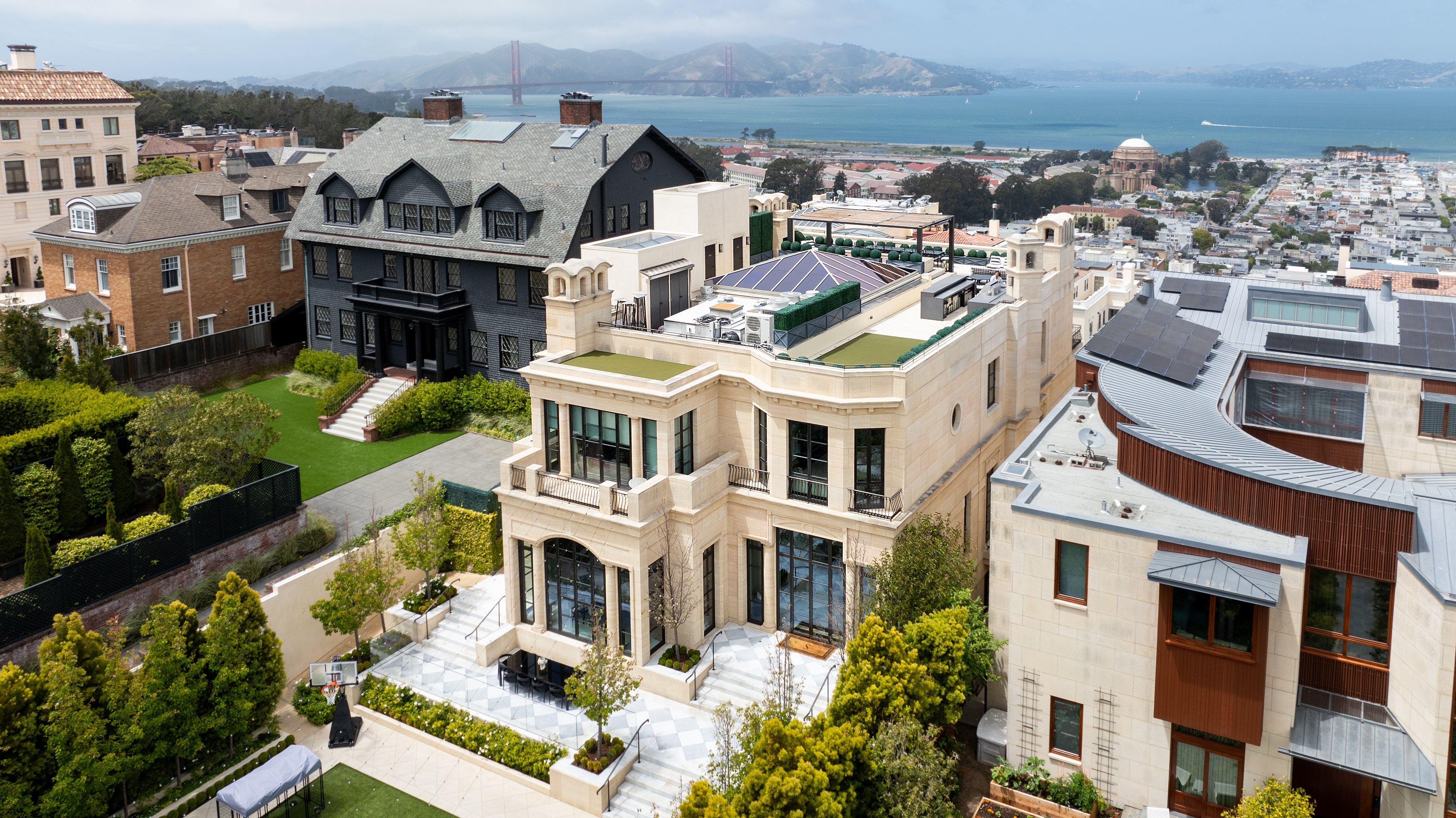 A luxurious neighborhood with elegant houses and manicured gardens. In the background, there's a scenic view of a bay with hills and a red bridge.