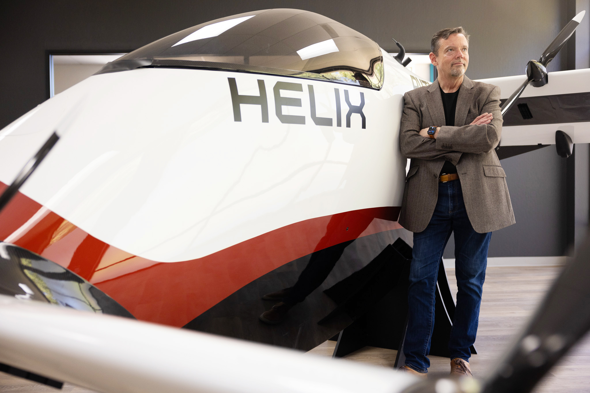 A man in a blazer and jeans stands with arms crossed next to a sleek, futuristic vehicle labeled &quot;HELIX,&quot; which has a glossy white and red exterior with a tinted canopy.