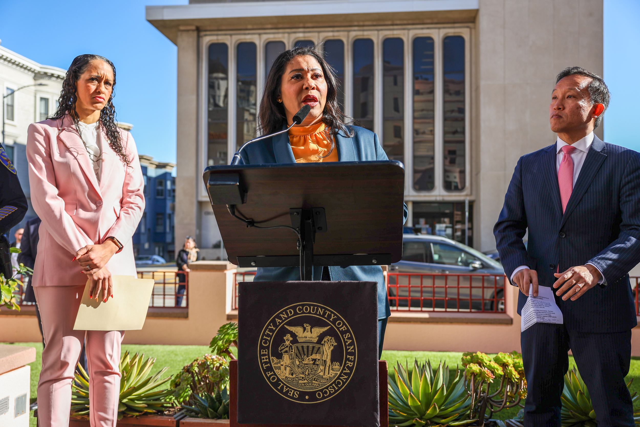 Three people in formal attire stand outdoors near a podium, which is adorned with a seal reading "City and County of San Francisco." One person speaks into a microphone.