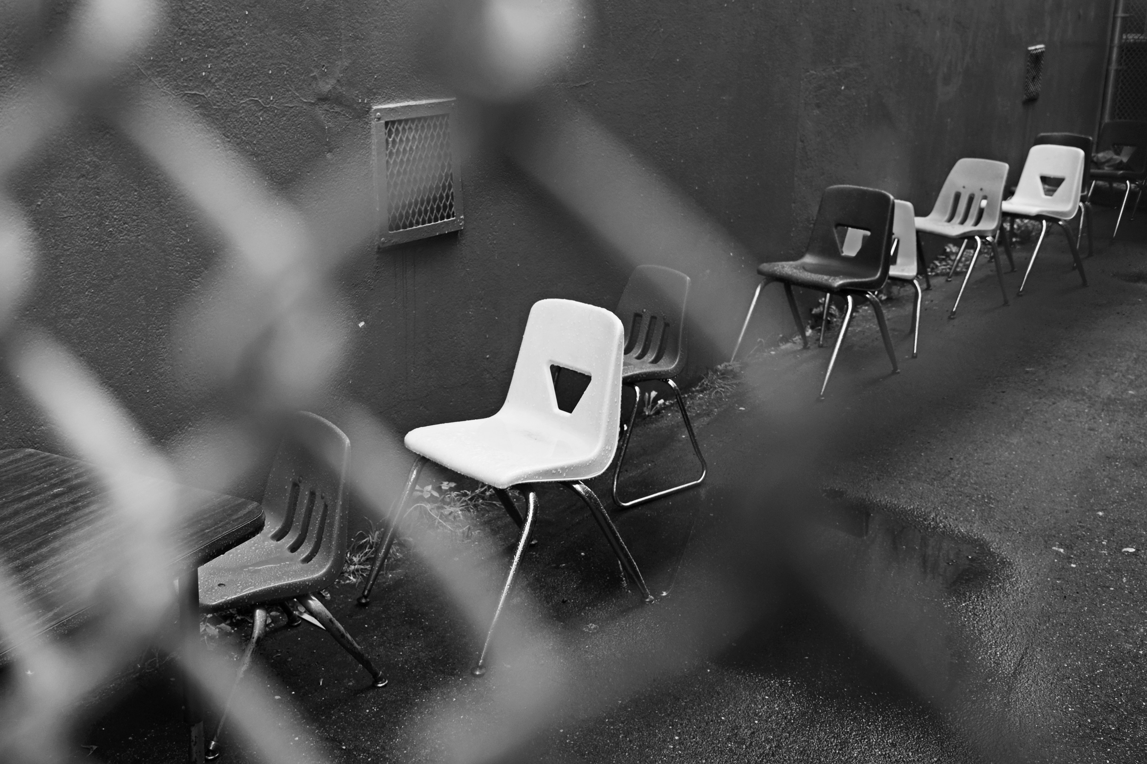 A row of mismatched plastic chairs, some white, black, and dark, is lined up against a wall on a wet, narrow alley. The image appears to be taken through a fence.