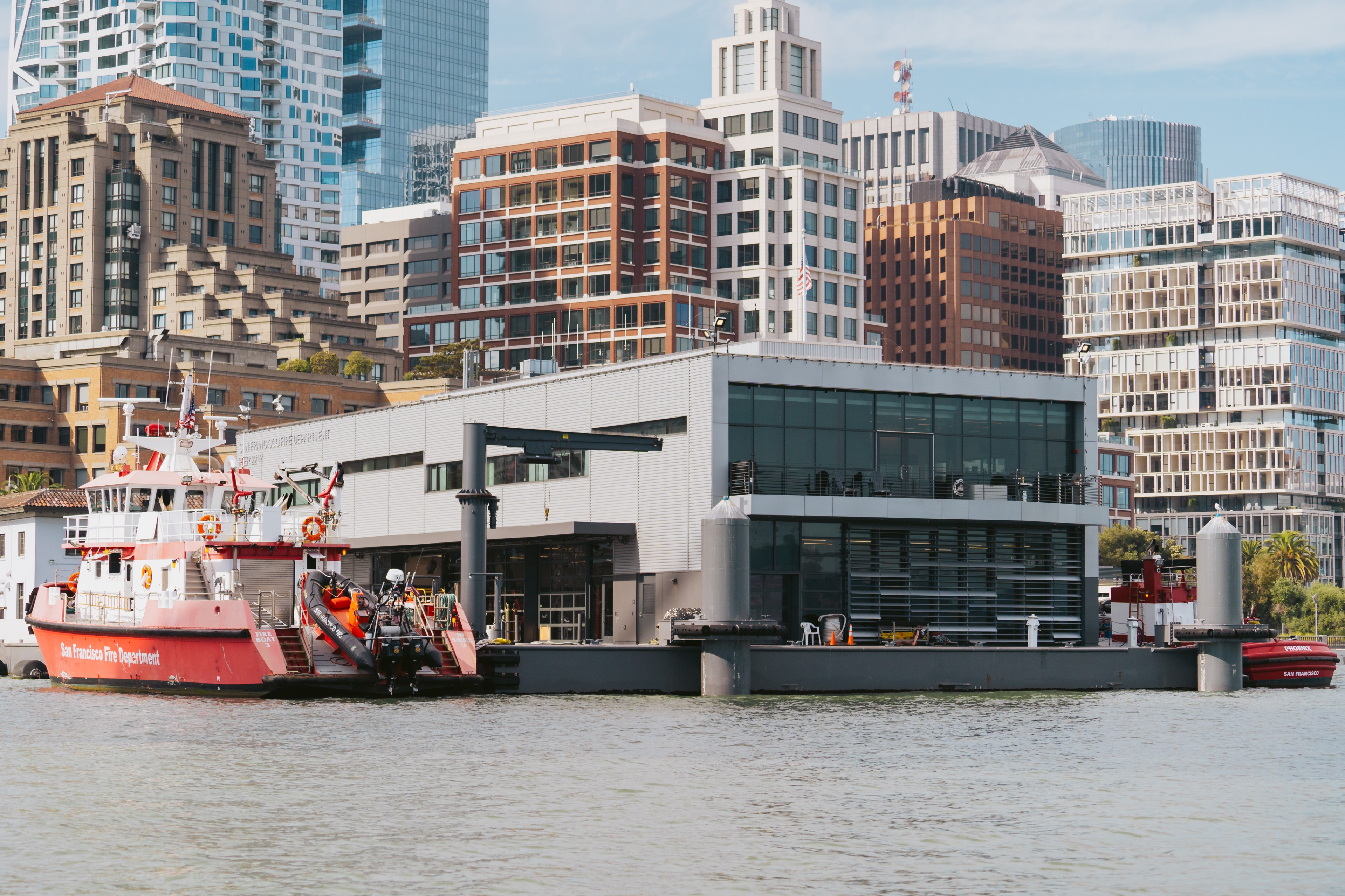A red and white fire department boat is docked near a modern, multi-story building along a waterfront, with various taller buildings in the background.