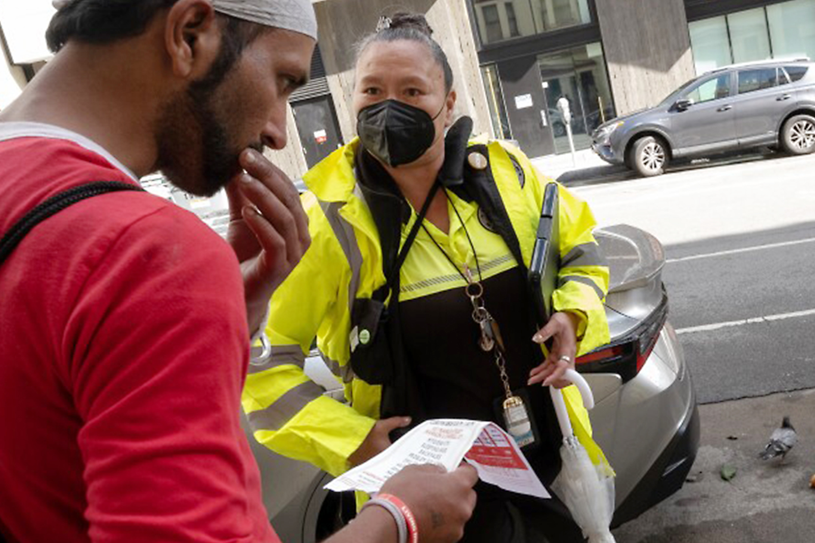 A person in a bright yellow safety jacket and black mask speaks with another person in a red shirt on a city street. They're discussing a document the second person is holding.