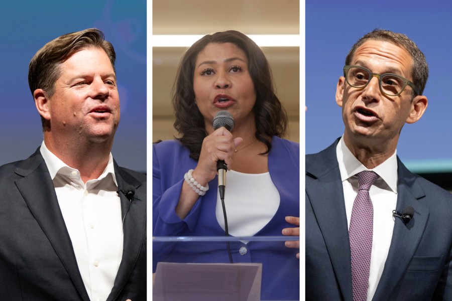 The image features three people. On the left, a man in a dark suit is speaking. In the middle, a woman in a blue blazer holds a microphone. On the right, a man in glasses and a suit is talking.