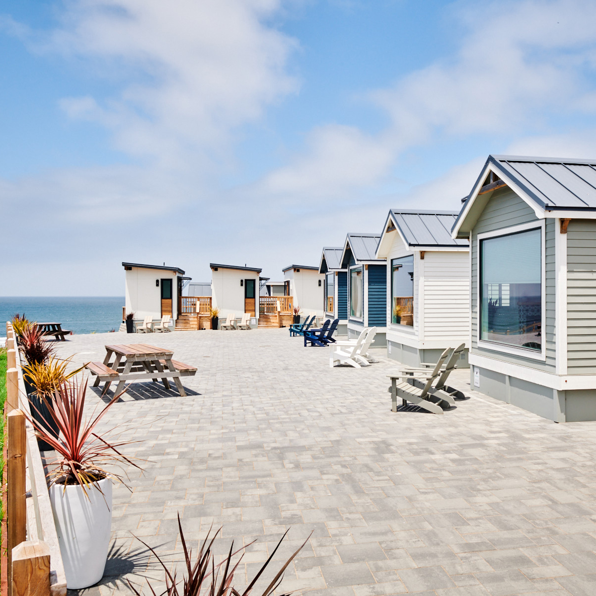 The image shows a row of modern, small coastal cabins with large windows and gray roofs, facing the ocean. There are deck chairs, picnic tables, and planters on a paved patio.