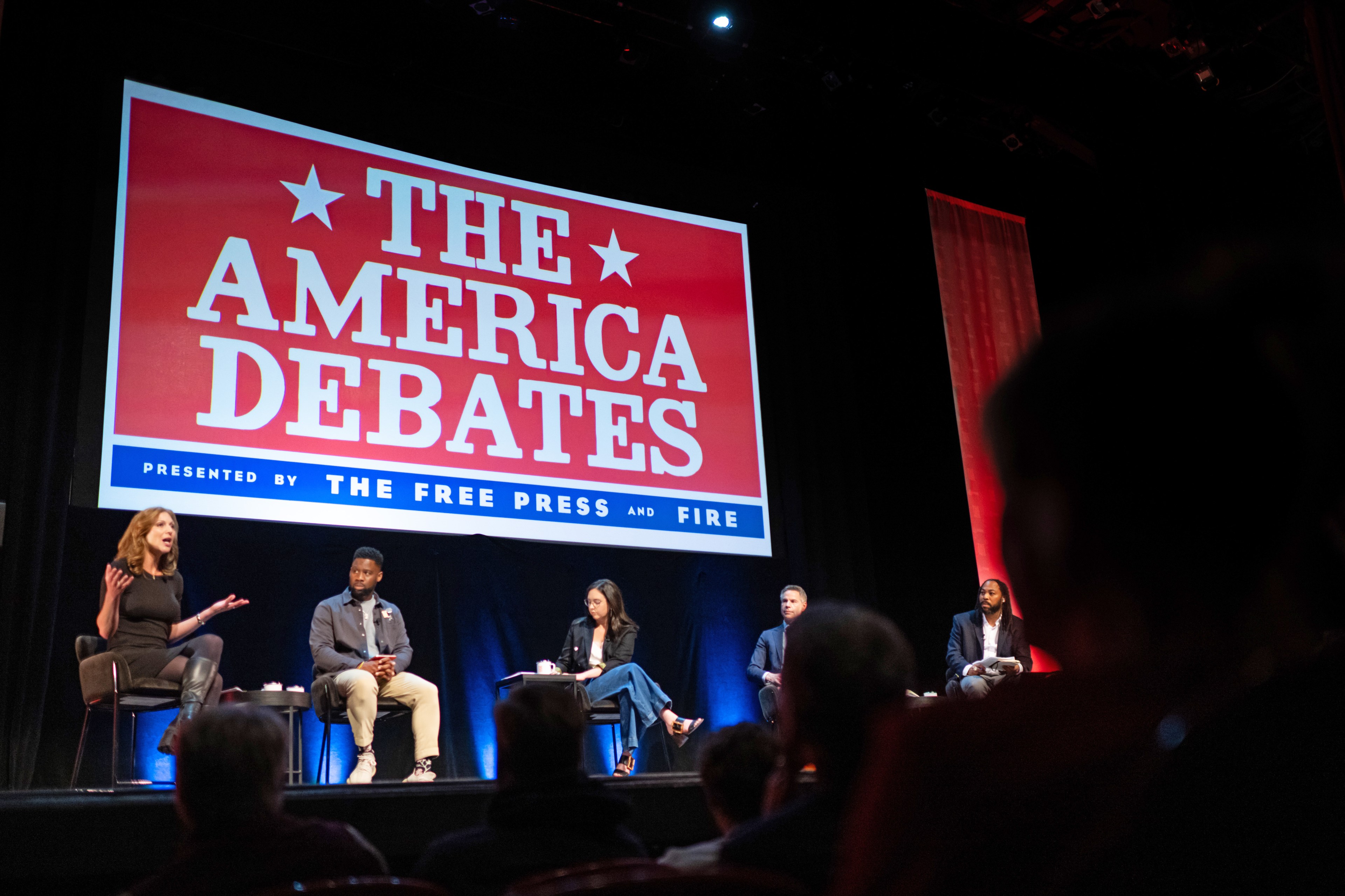 Five individuals are seated on stage under a large sign reading "The America Debates," hosted by The Free Press and FIRE, engaging in a discussion or debate.