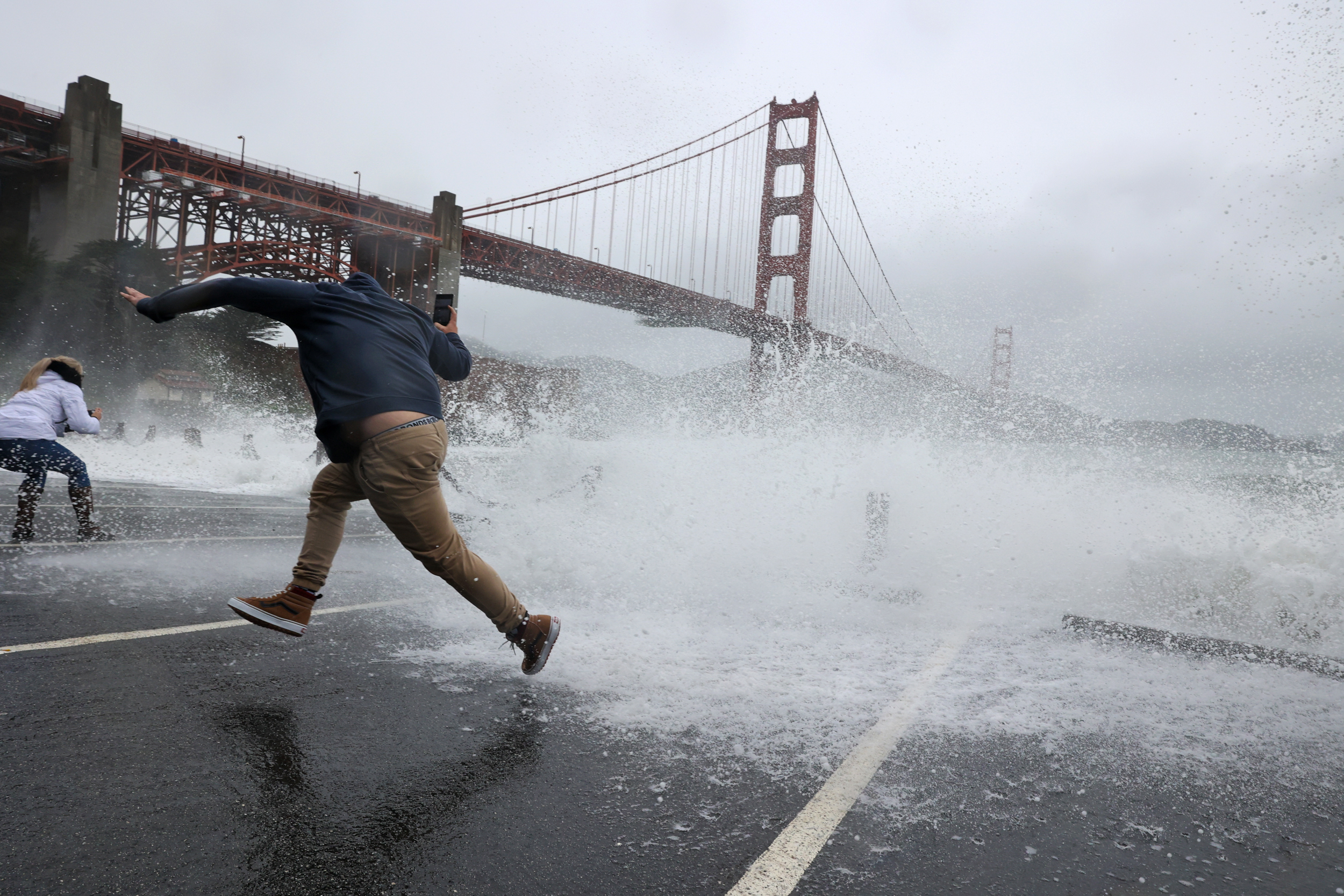 In San Franciso, beneath the Golden Gate Bridge, a man jumps over a wave that hits the pavement.