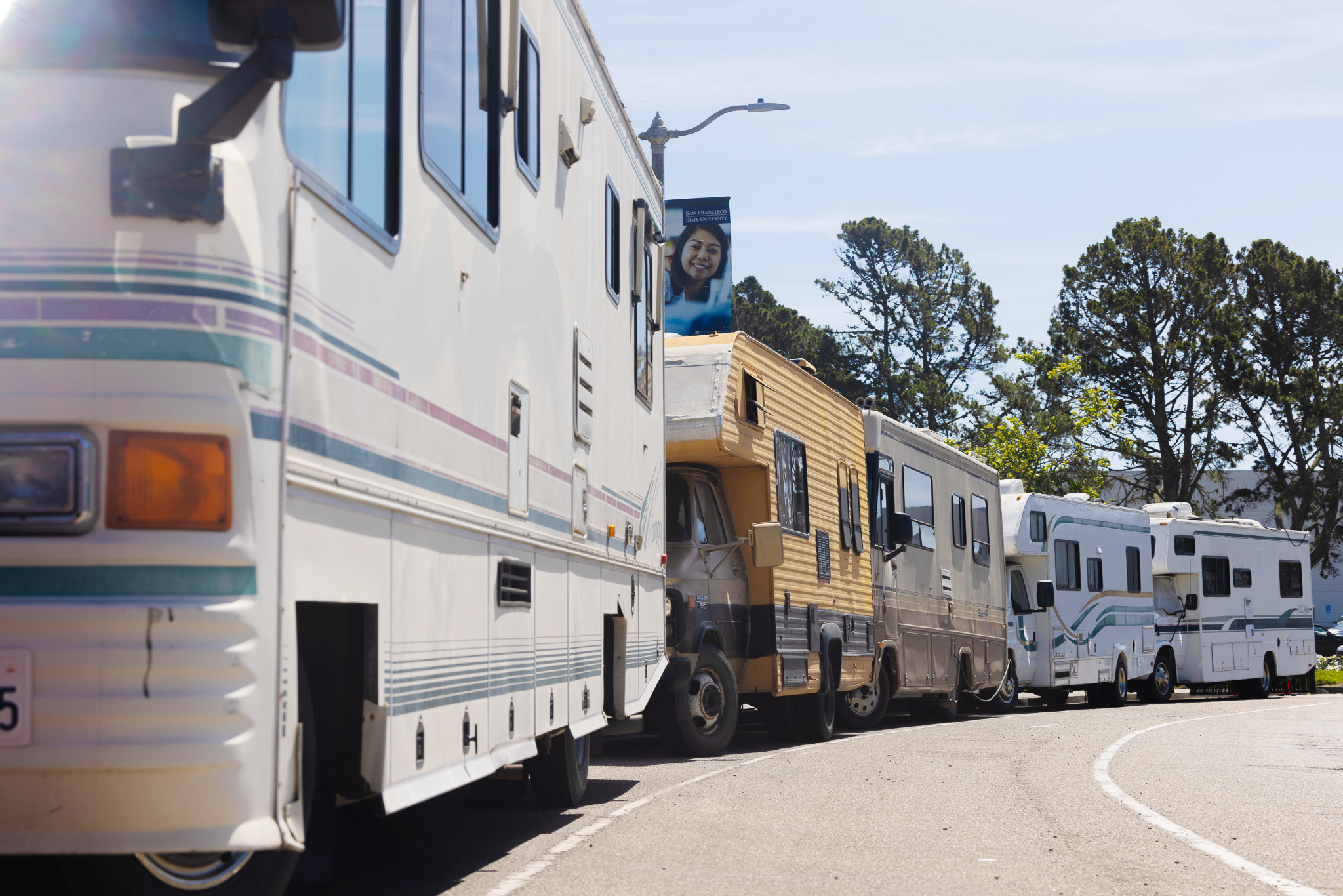 Several RVs parked closely along a roadside under a sunny sky, with trees and a banner featuring a smiling woman in the background.