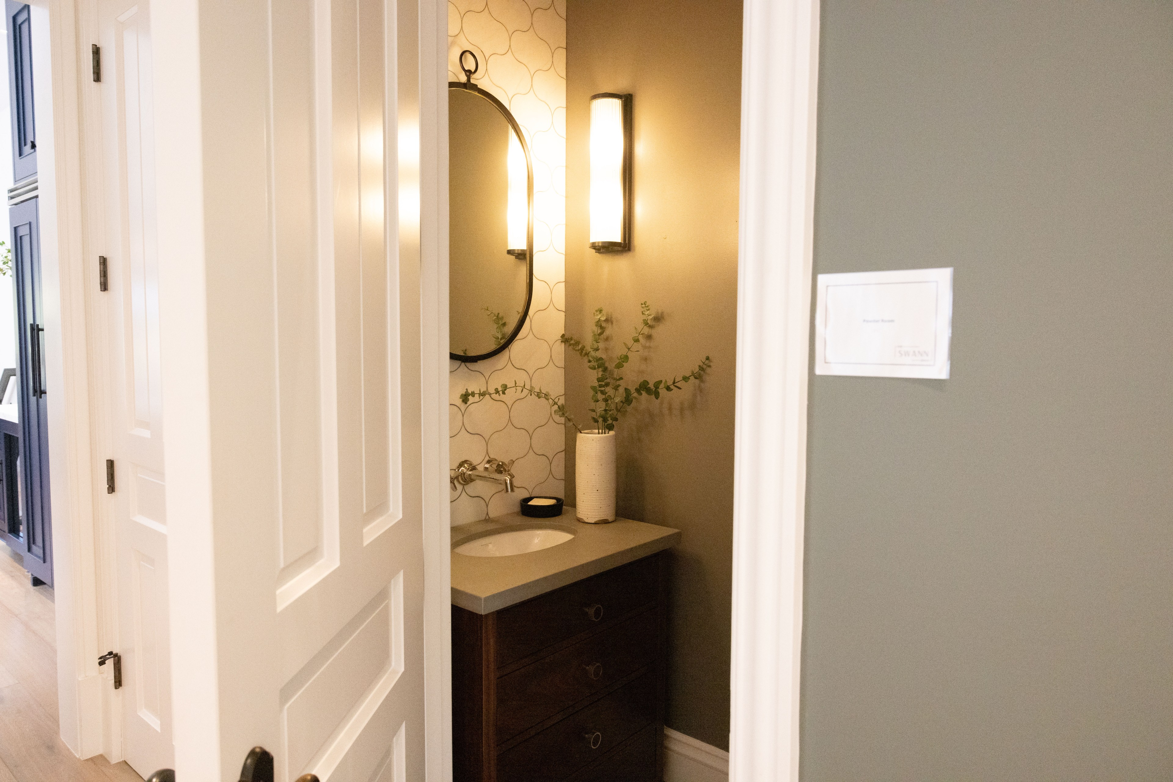The image shows a small bathroom with a vanity, round mirror, wall sconce, and modern decor, including a vase with greenery, visible through a white door.