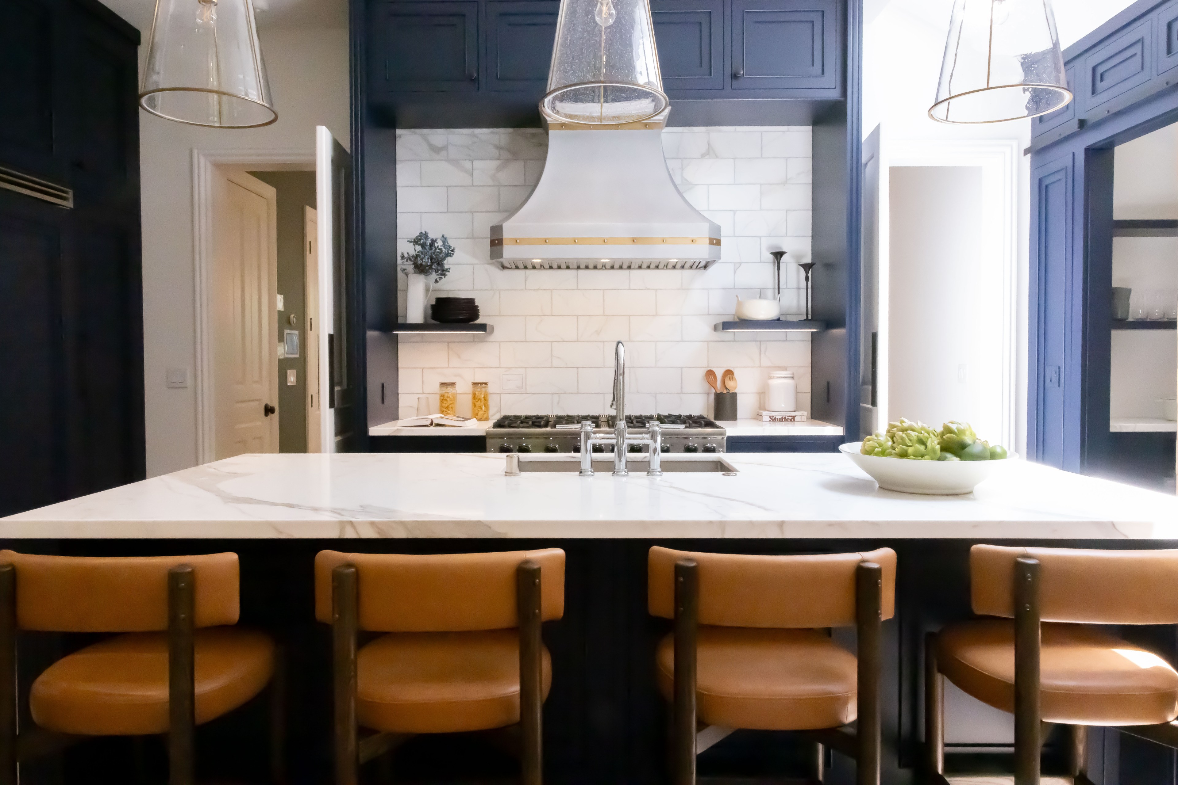 The image shows a modern kitchen with a large white marble island, four tan leather barstools, navy cabinets, stainless steel appliances, and hanging glass pendant lights.