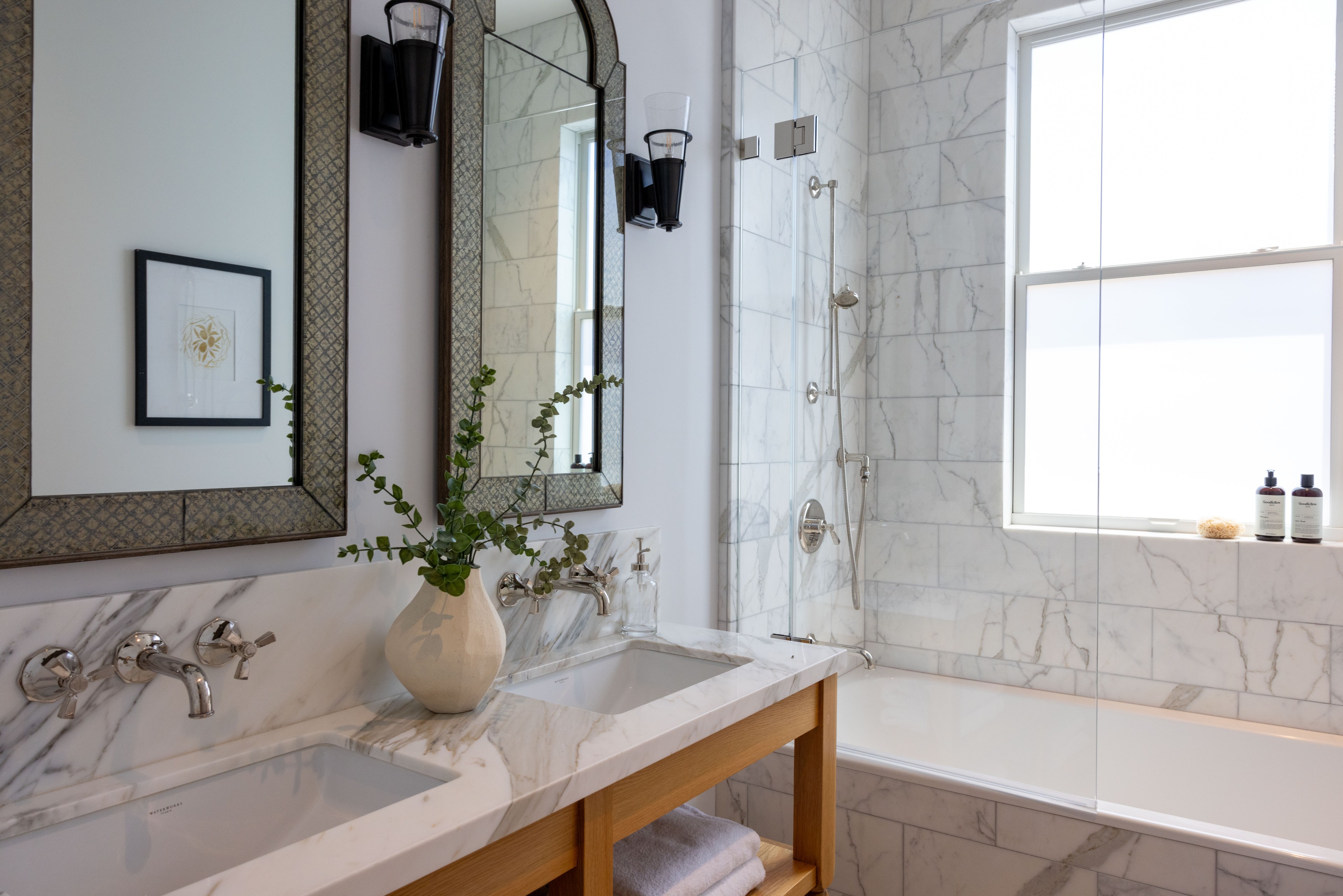 The image shows a stylish bathroom with white marble countertops and walls, dual sinks, decorative mirrors above, a white vase with green leaves, and a bathtub with a shower.