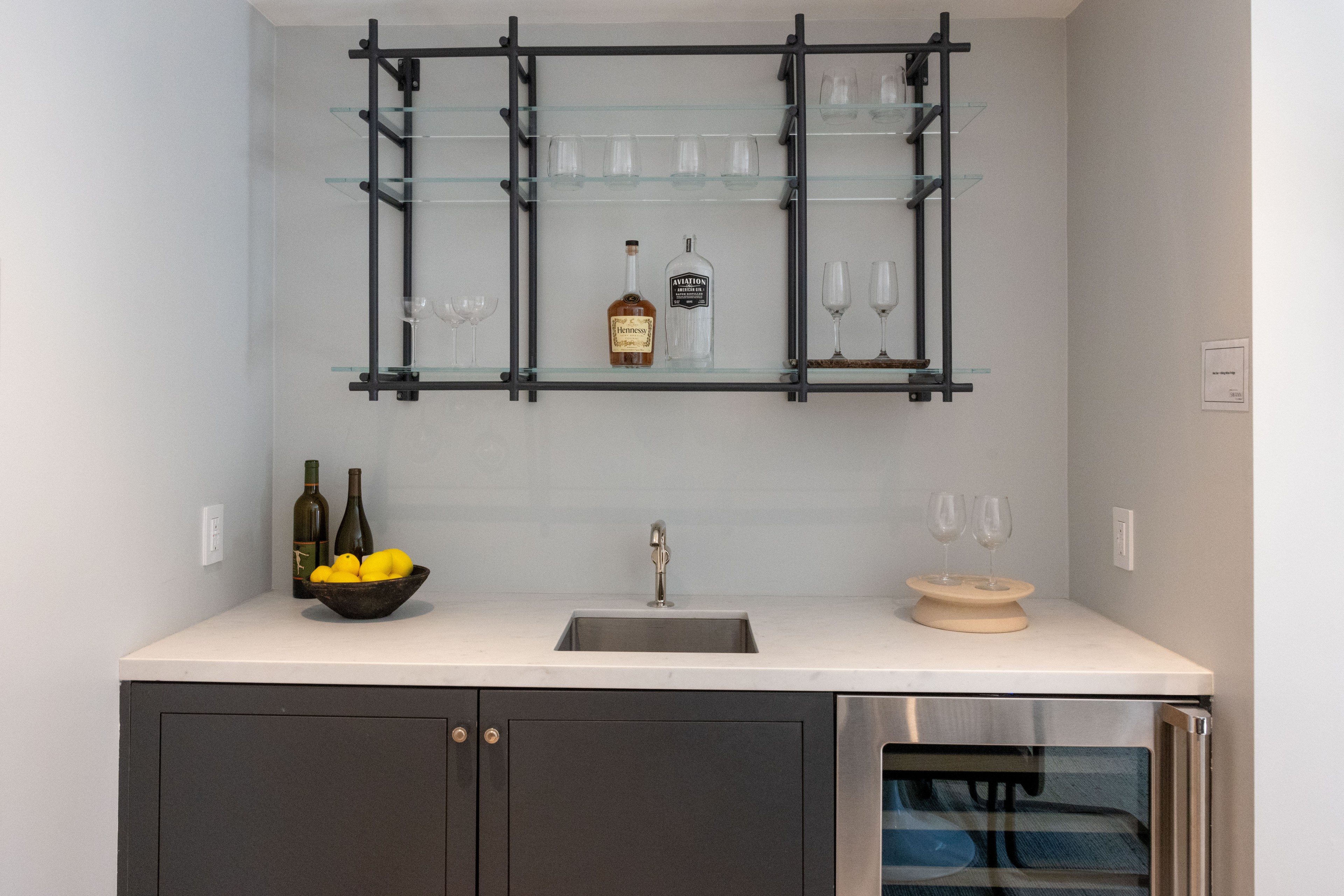 A compact home bar with a marble countertop, a small sink, and dark lower cabinets. Shelves above hold glasses, a bottle of Hennessy, a gin bottle, and lemons.