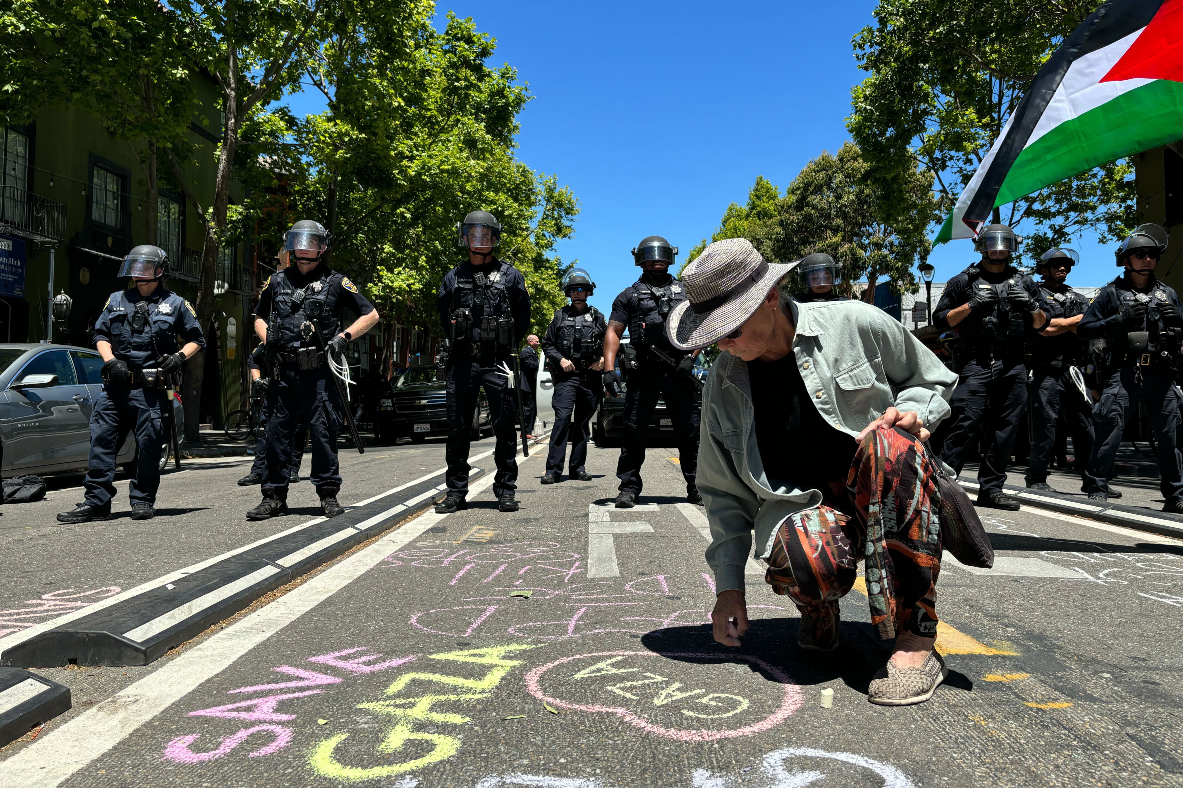 A person is writing &quot;SAVE GAZA&quot; on the street with chalk, while a line of police officers in riot gear stands behind them. A Palestinian flag is visible on the right.