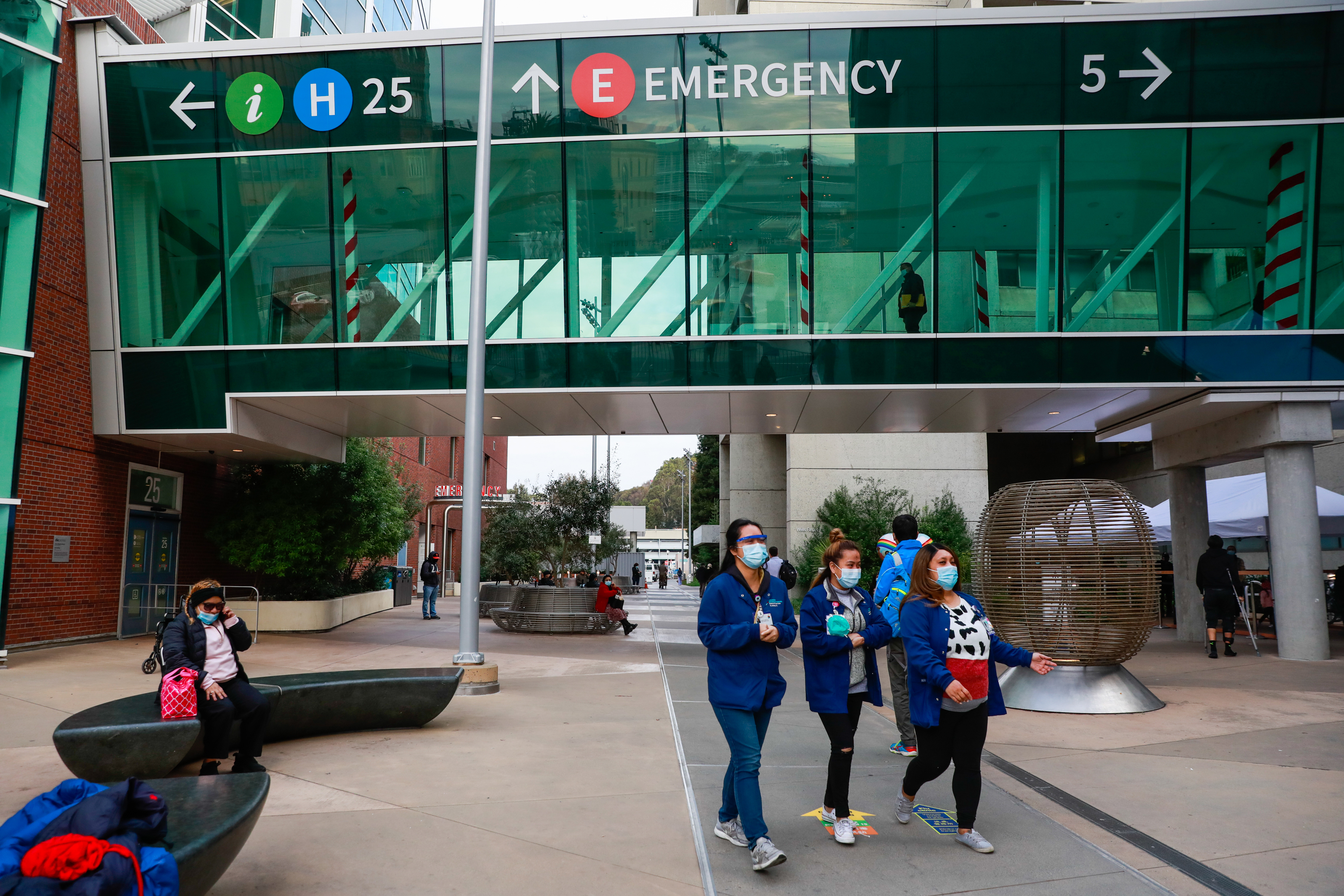 Four people wearing masks walk under a glass bridge with signs overhead pointing to information, hospital, and emergency departments.