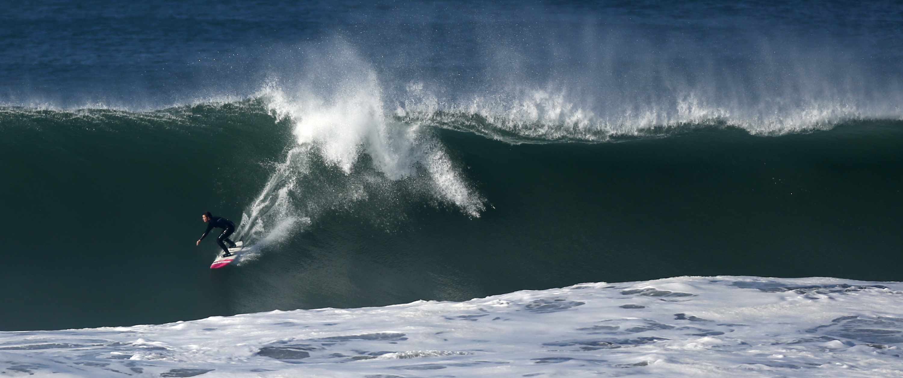 A surfer rides a large, powerful wave with white foam at the base, in the deep blue ocean, creating a dynamic and intense scene full of motion.