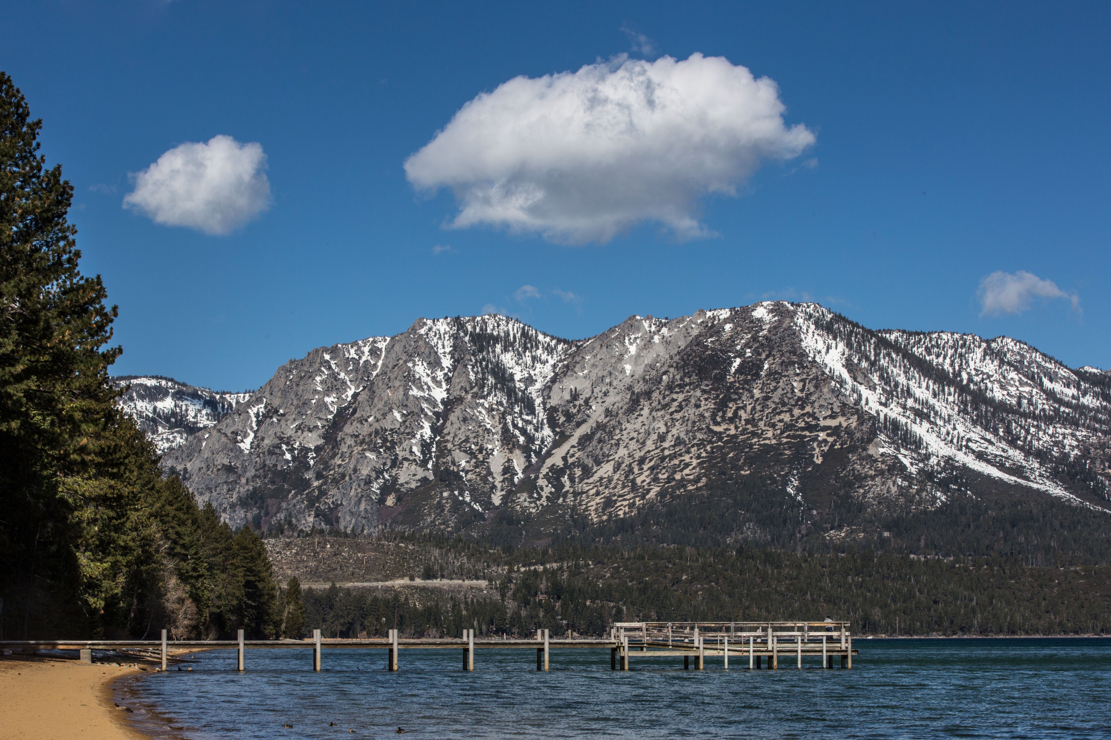 This image shows a serene lake with a sandy shore, a wooden pier, and snow-capped mountains in the background under a clear blue sky with a few fluffy clouds.