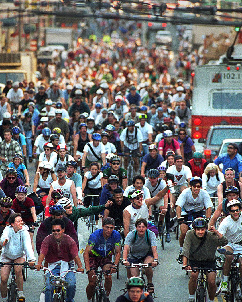 A large crowd of cyclists, wearing helmets and casual attire, fill the street. Bicycles extend as far as the eye can see, with an emergency vehicle on the right.