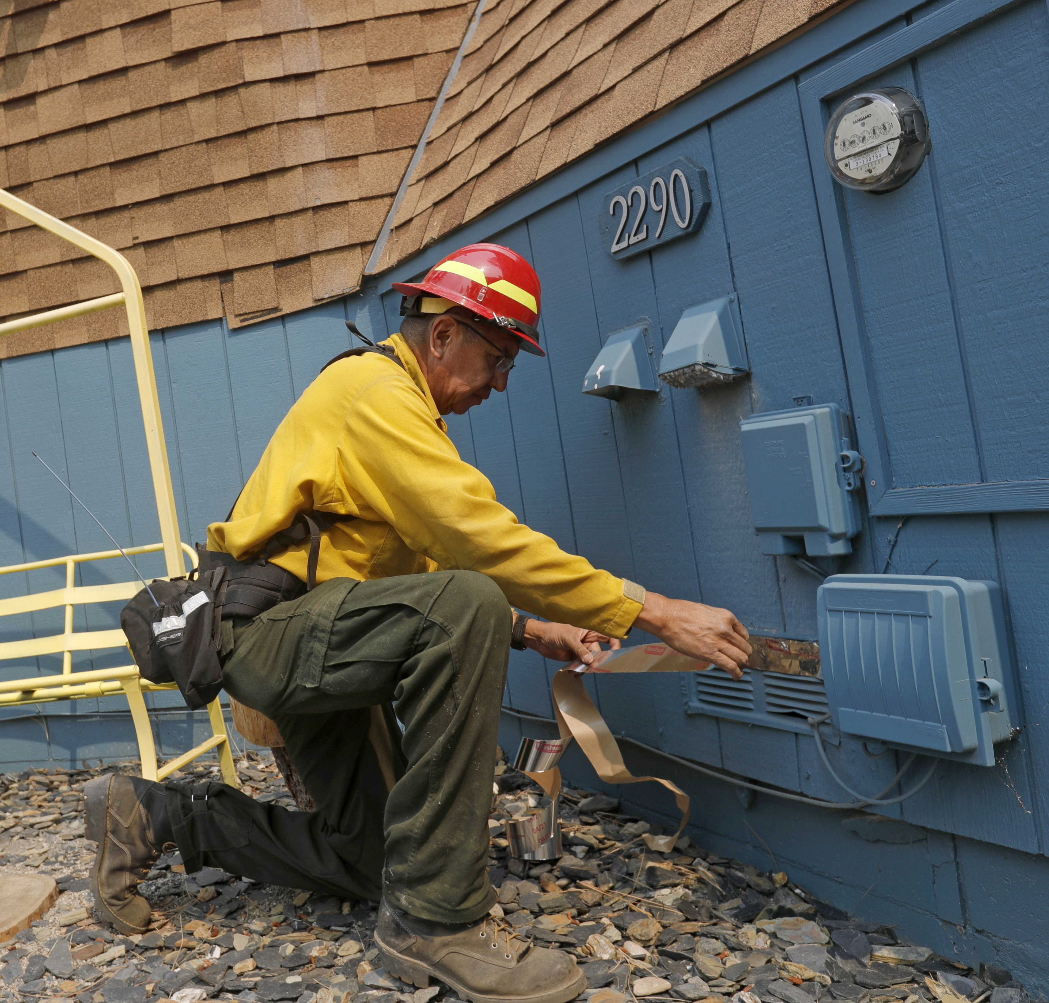 A utility worker, wearing a red helmet and yellow shirt, kneels beside a blue house numbered 2290, working on an electrical box with duct tape.