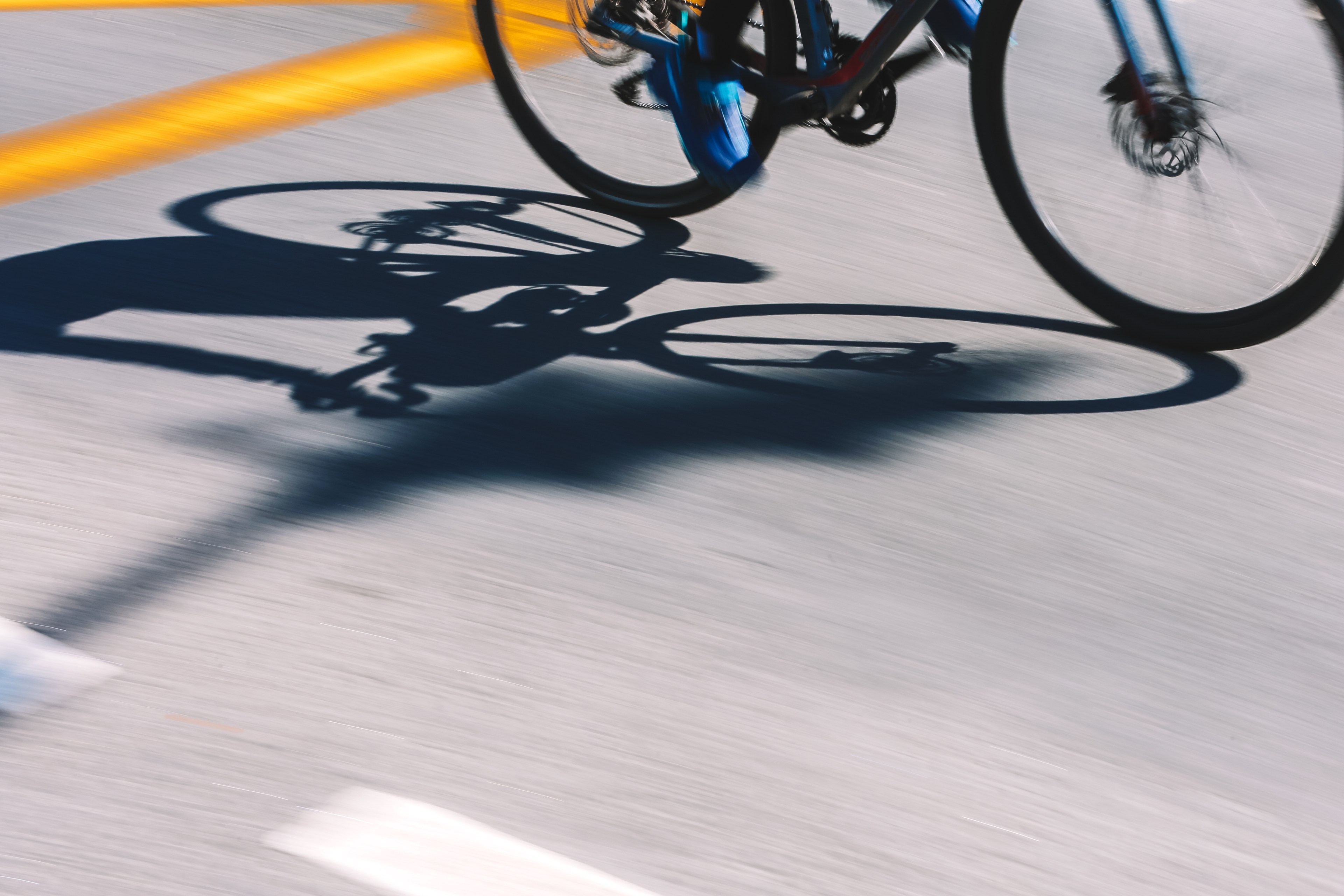 The image shows a blurred close-up of a bicycle in motion on a road, casting a shadow. The shadow and bike wheels create dynamic lines on the surface.
