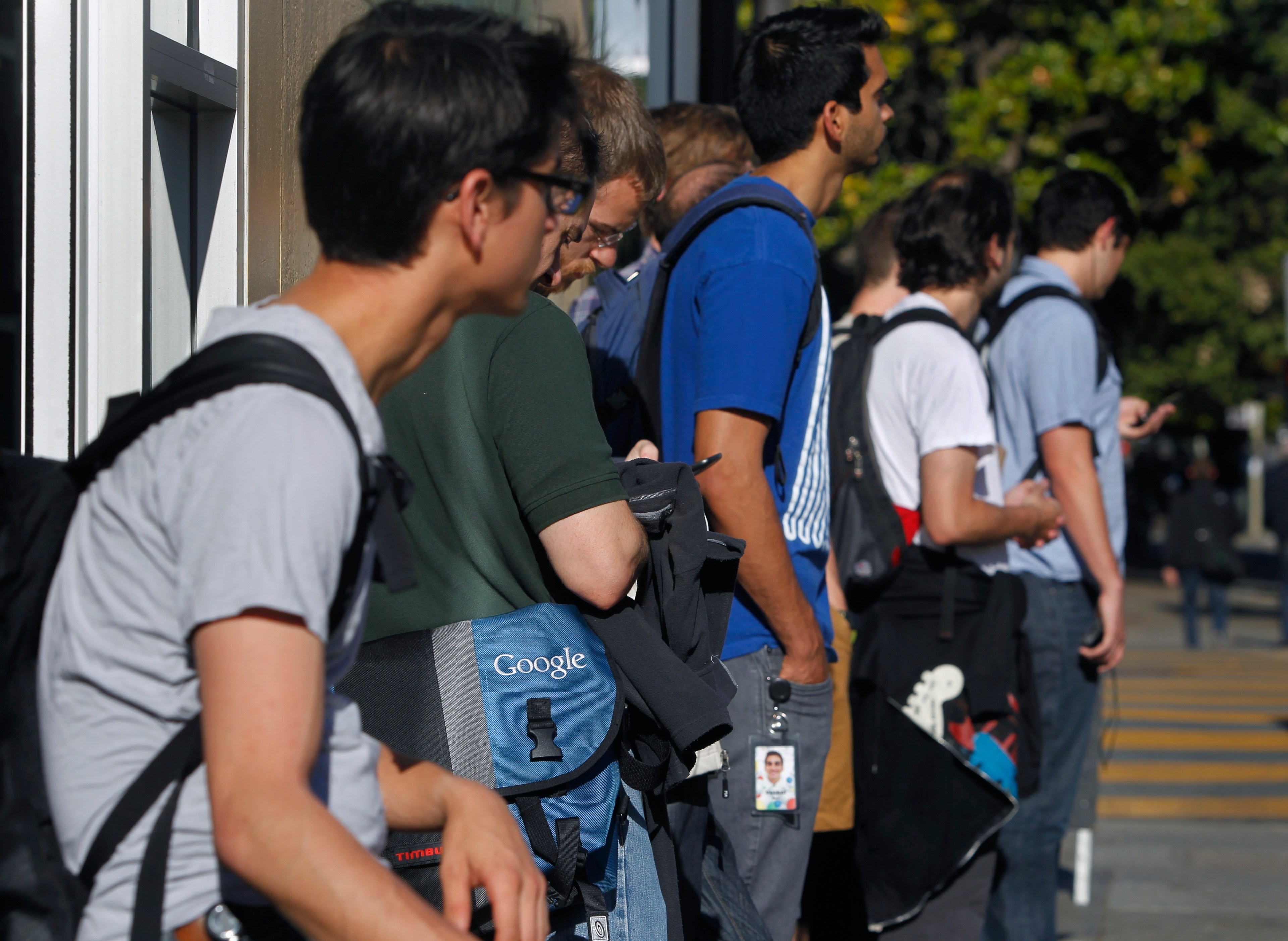 A group of people, mostly young men with backpacks and messenger bags, are standing in line outdoors. One visible bag reads &quot;Google.&quot;