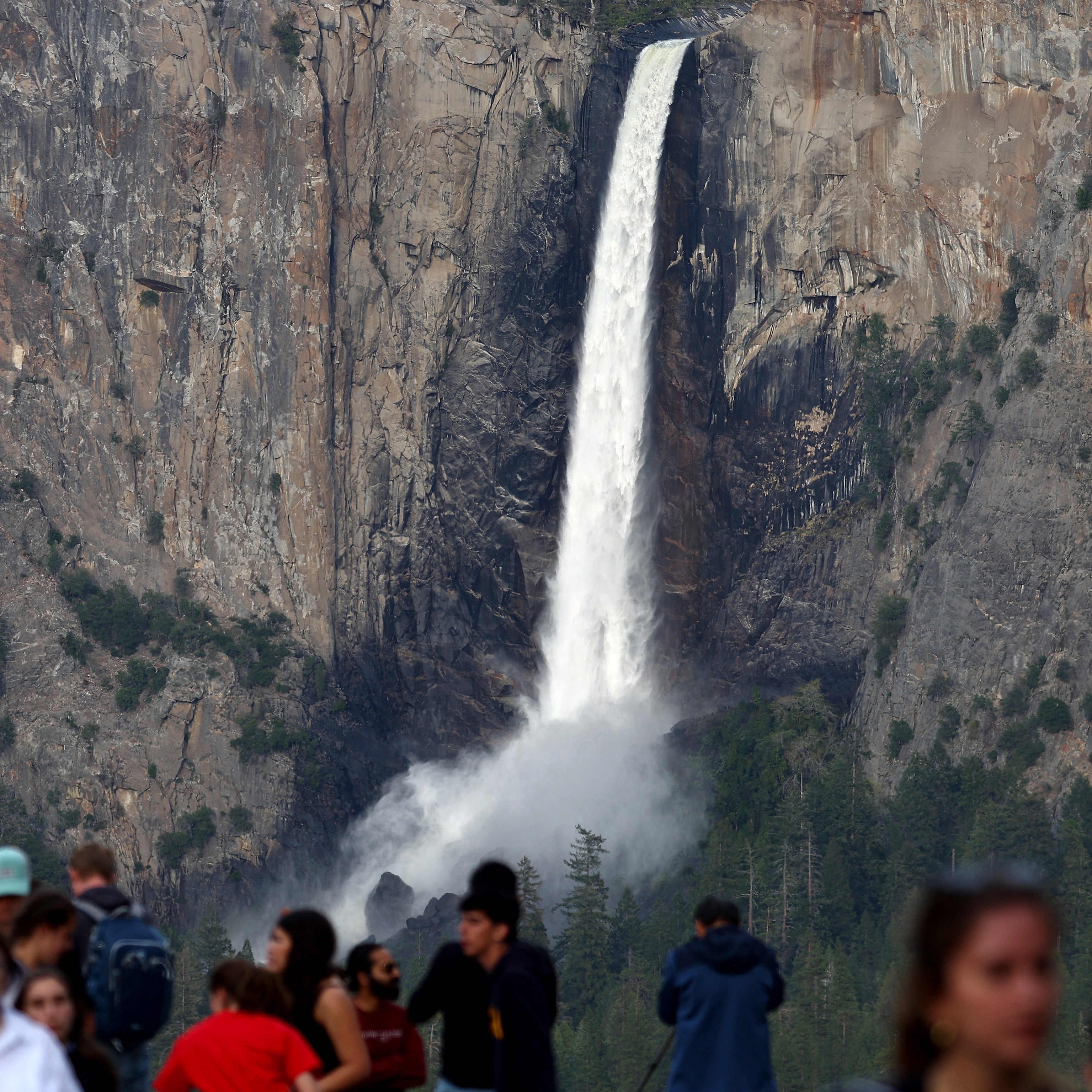 A tall waterfall cascades down a rugged cliffside with mist rising at its base. In the foreground, a crowd of people gathers, partially blurred, observing the scenery.