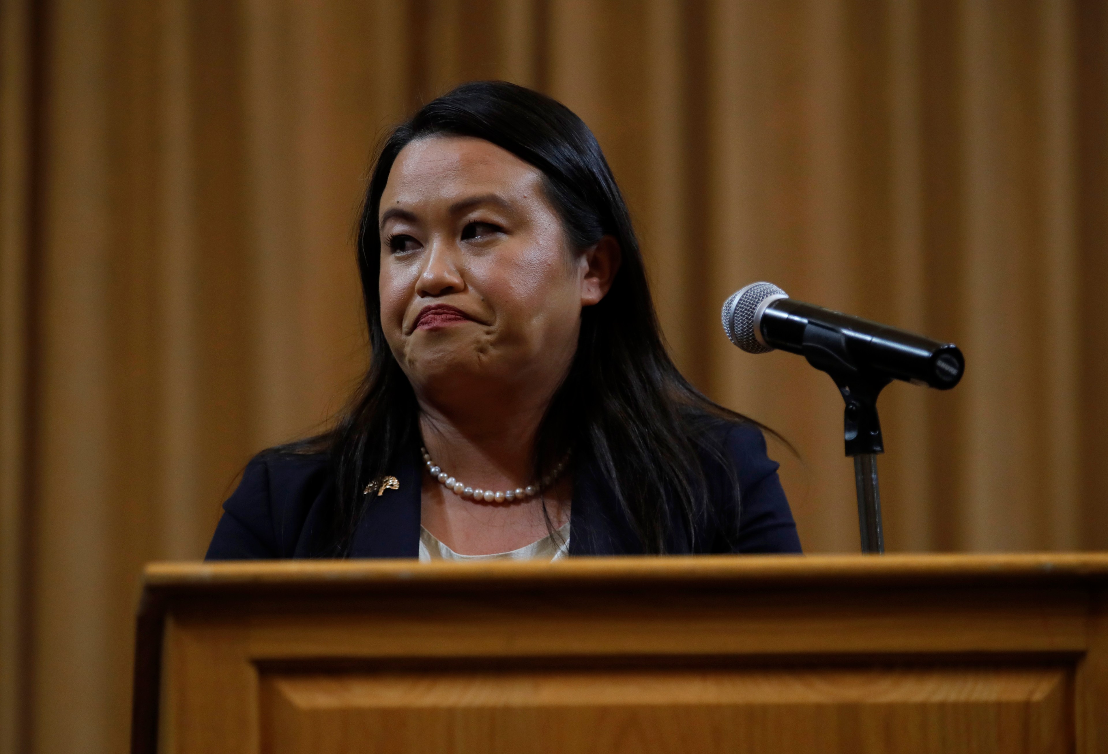 A woman stands at a podium with a microphone, wearing a dark blazer and pearl necklace, against a plain curtain backdrop. Her expression appears thoughtful.