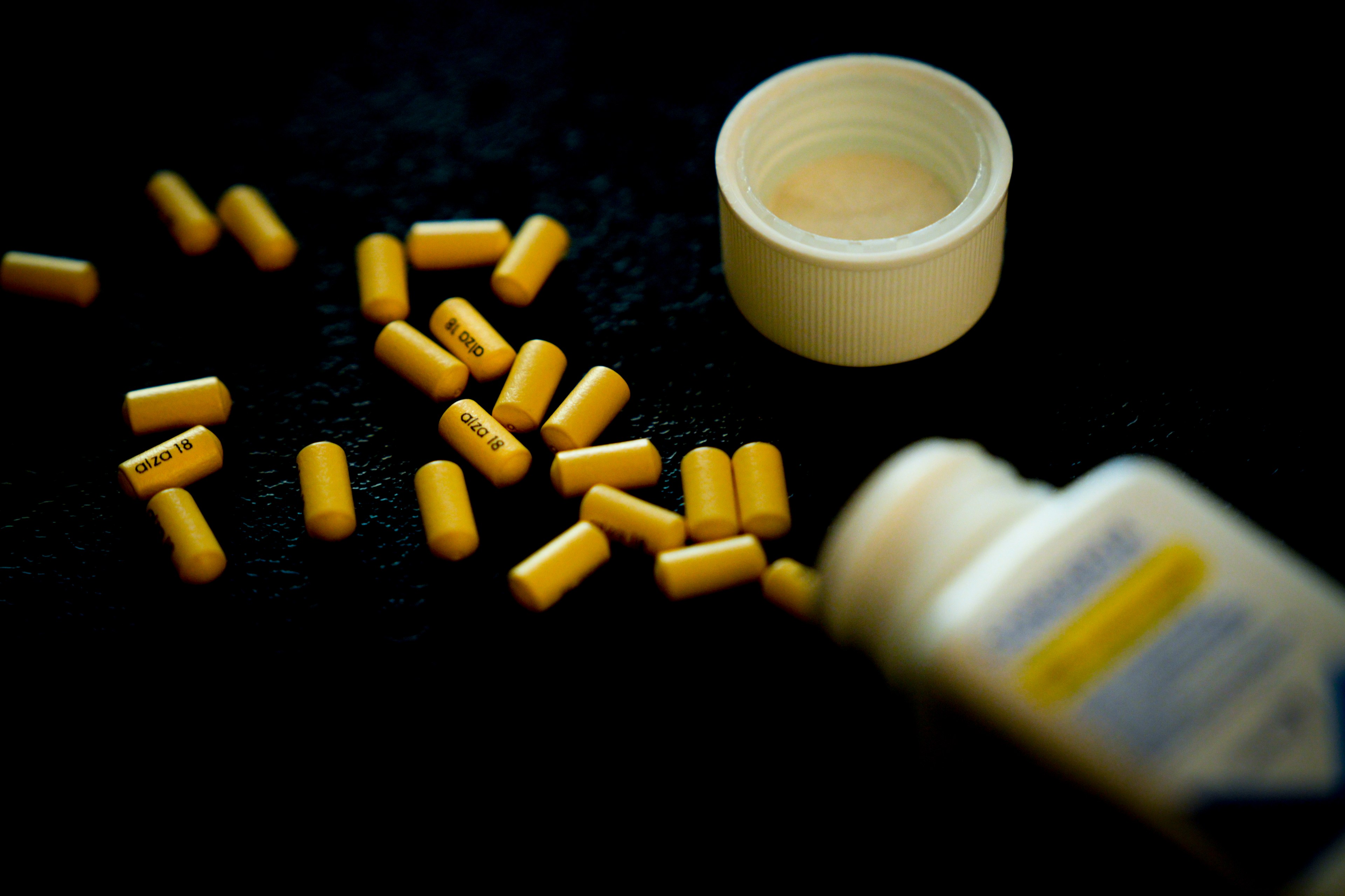 Yellow capsules have spilled onto a dark surface from a white, open pill bottle, which has its cap next to it. Some capsules are scattered around.