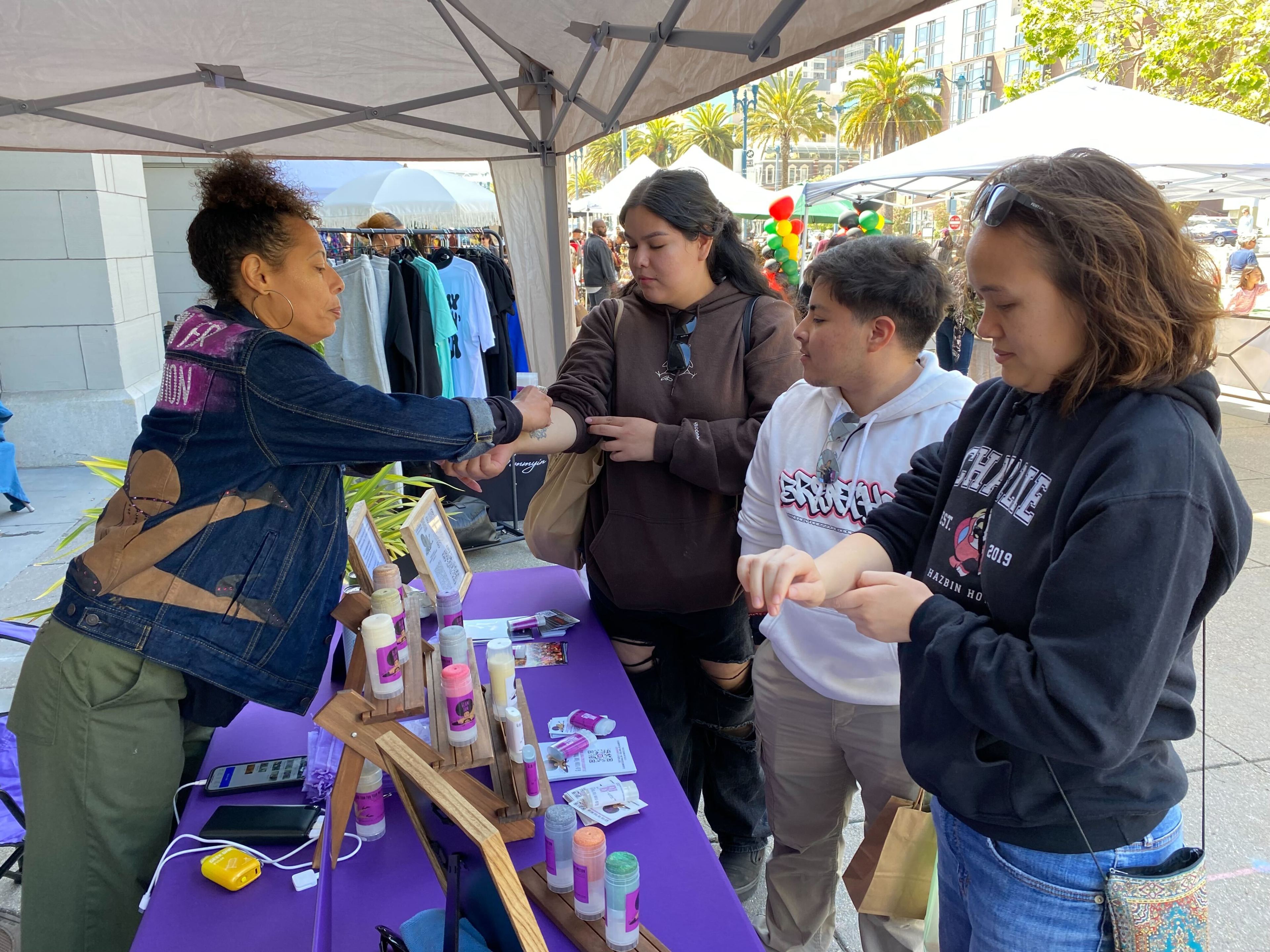 A woman at a booth applies lotion to three people’s arms under a canopy at an outdoor market, with various skincare products displayed on the purple-covered table.