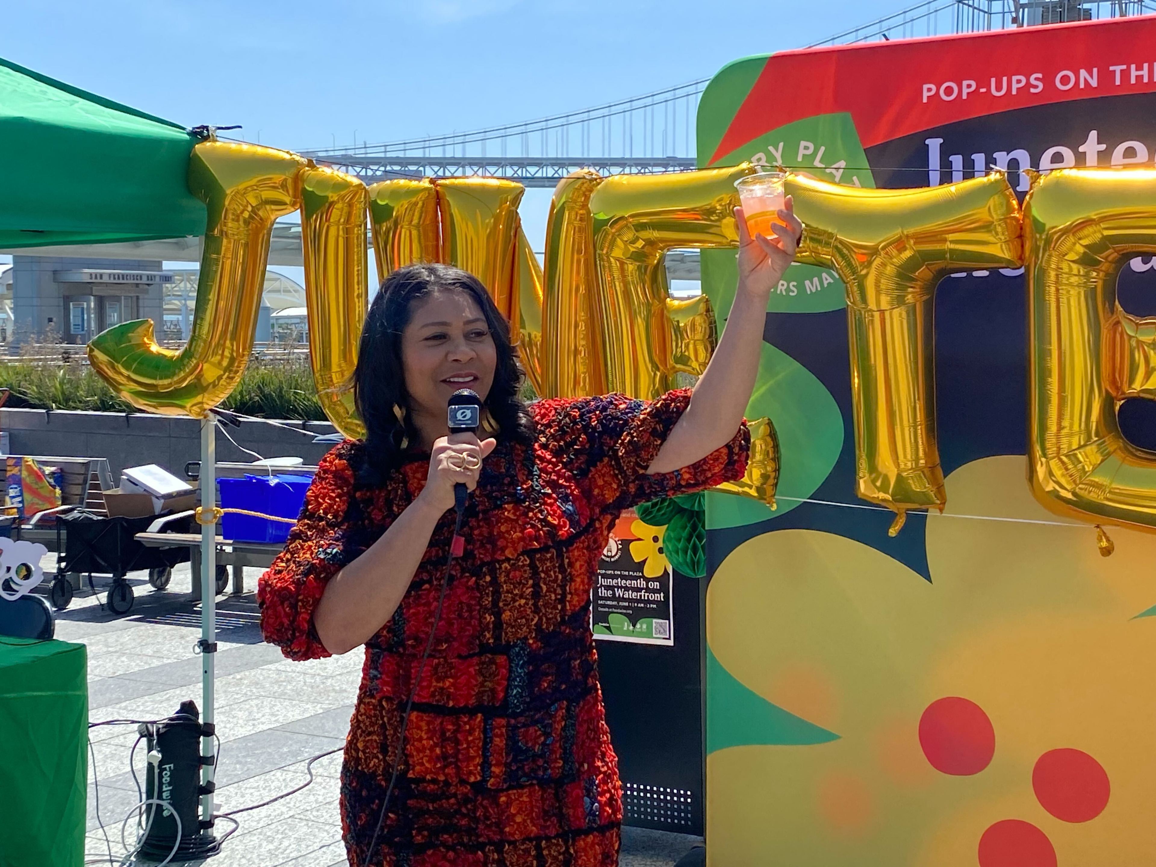 A woman in a patterned dress speaks into a microphone and raises a drink. Behind her, gold balloons spell "JUNETEENTH." A bridge and a green tent are visible in the background.