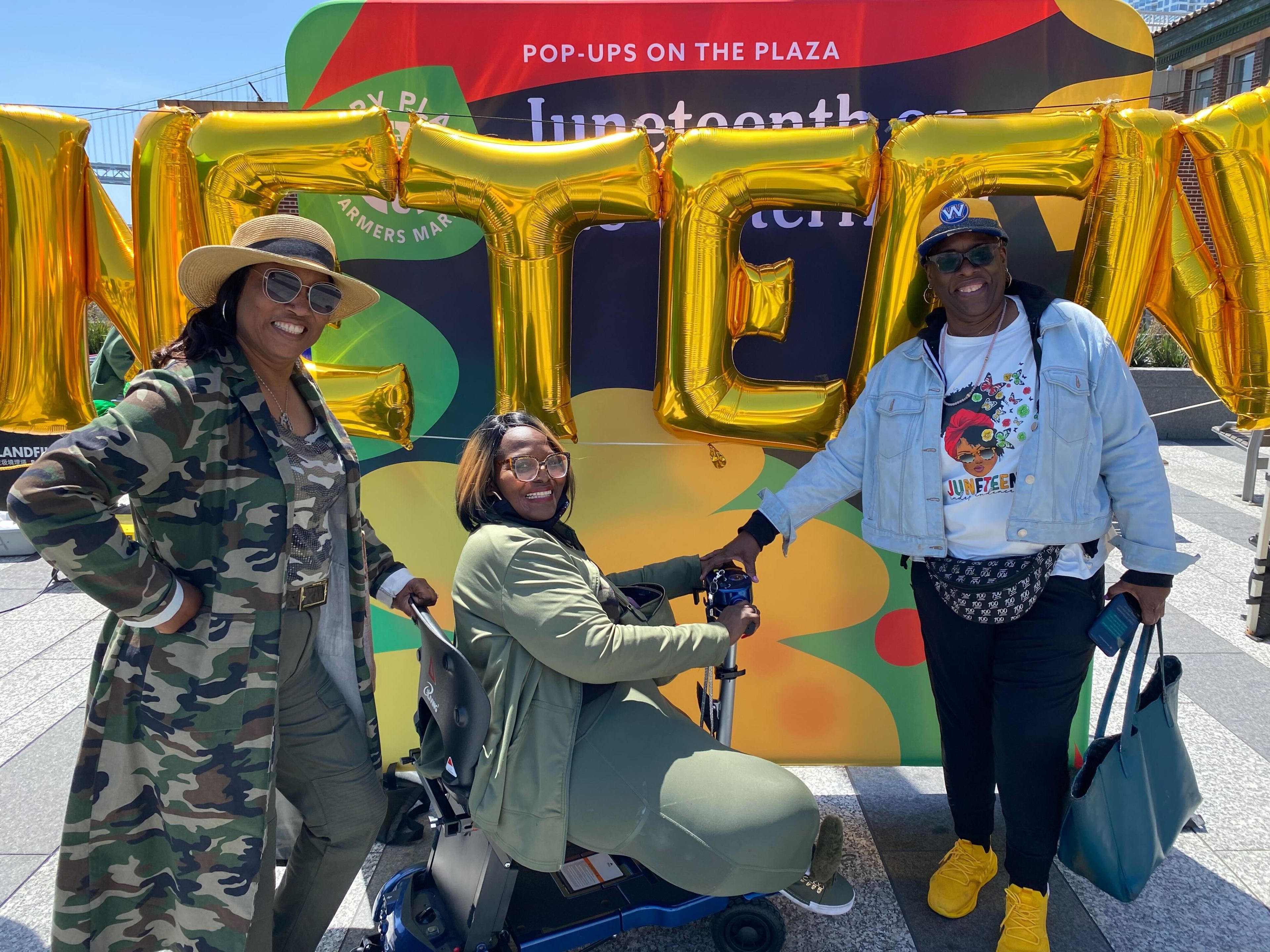 Three women smile in front of large gold balloons spelling &quot;JUNETEENTH,&quot; with one seated on a mobility scooter. They are outdoors, dressed in casual attire.