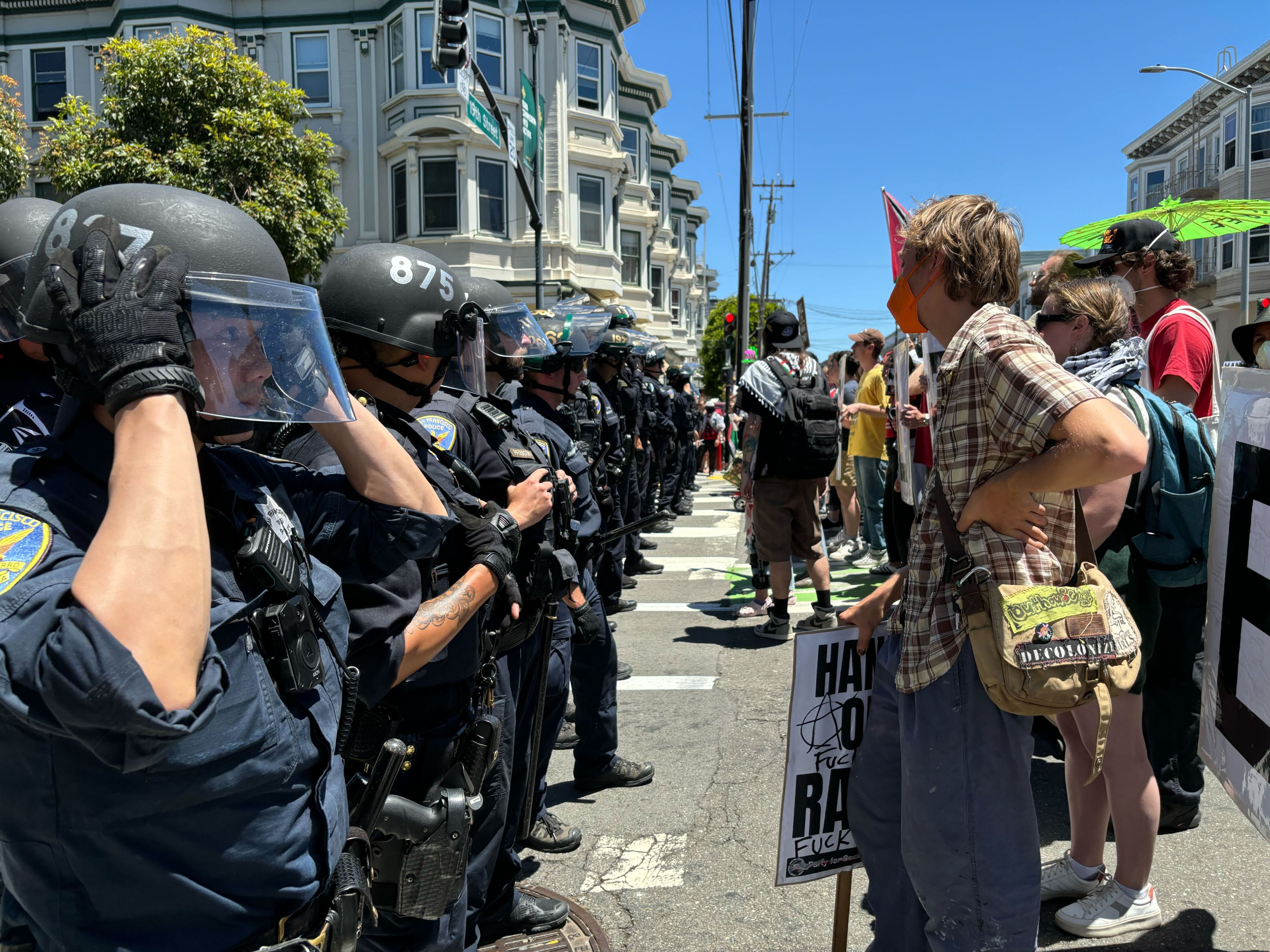 A line of police officers in riot gear face off against protesters in a city street. The scene includes both officers and civilians holding signs in an urban setting.