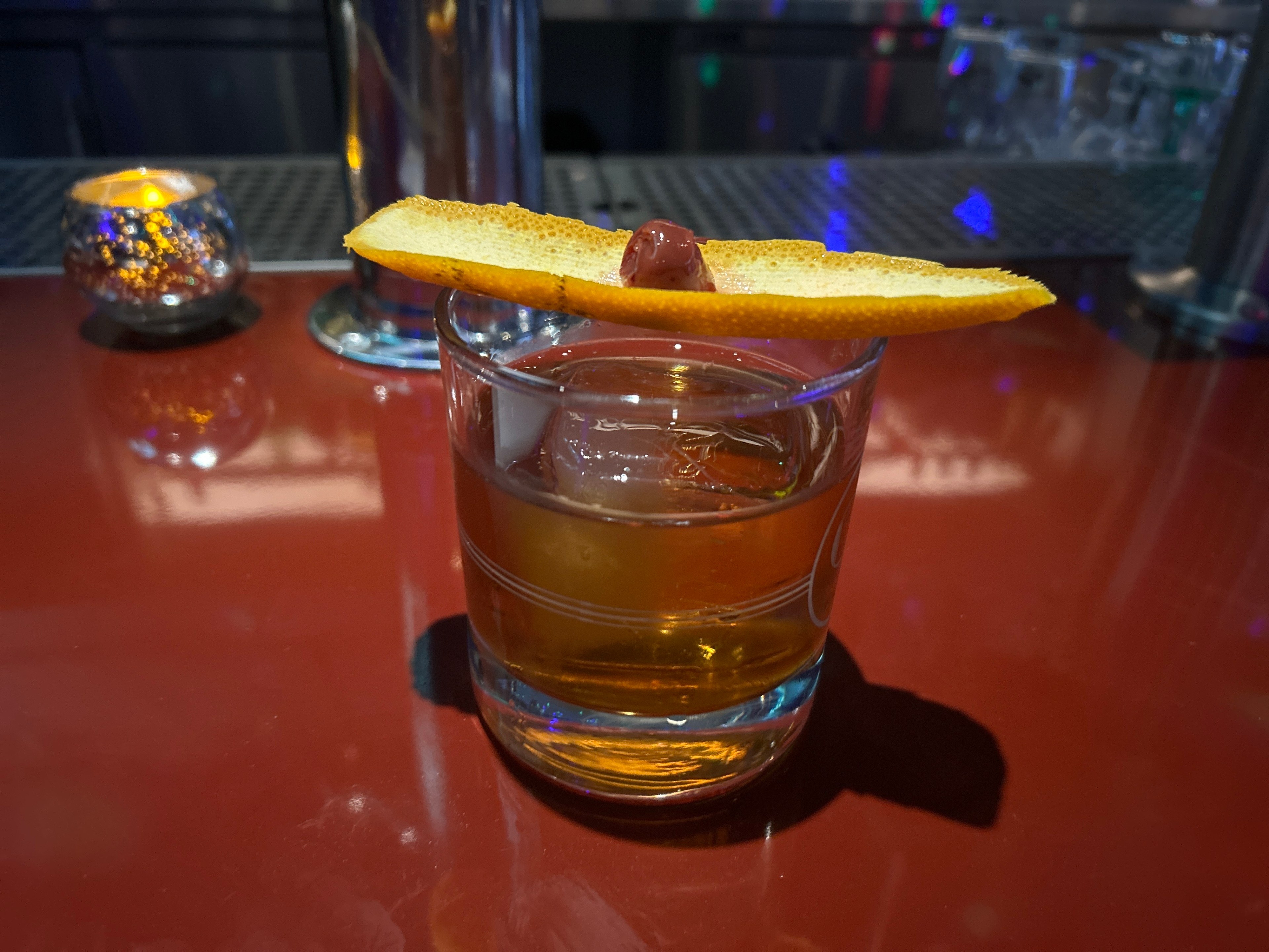 A brown cocktail on the rocks garnished with an orange peel sits on a red bar.